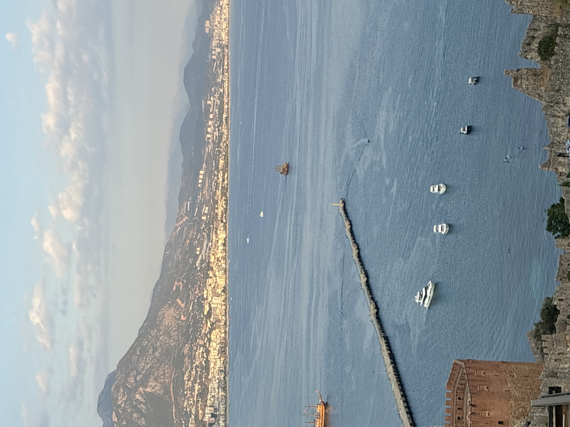 The image shows a scenic view of the coastline of Alanya, Turkey, with the Mediterranean Sea in the foreground and mountains in the background. The sun is setting, casting a warm glow on the landscape.
In the foreground, a section of Alanya Castle is visible, with its characteristic red brick walls and battlements. The sea is calm and blue, with several boats scattered across its surface. One of the boats is a large, wooden sailing ship, possibly a tourist boat. There is also a long pier extending out into the water, with a lighthouse at its end.
In the middle ground, the city of Alanya stretches along the coast, with its buildings and houses nestled at the foot of the mountains. The mountains themselves are covered in lush vegetation, and their peaks are shrouded in a light haze. The sky is a pale blue, with a few scattered clouds. The overall impression is one of a peaceful and picturesque coastal scene.