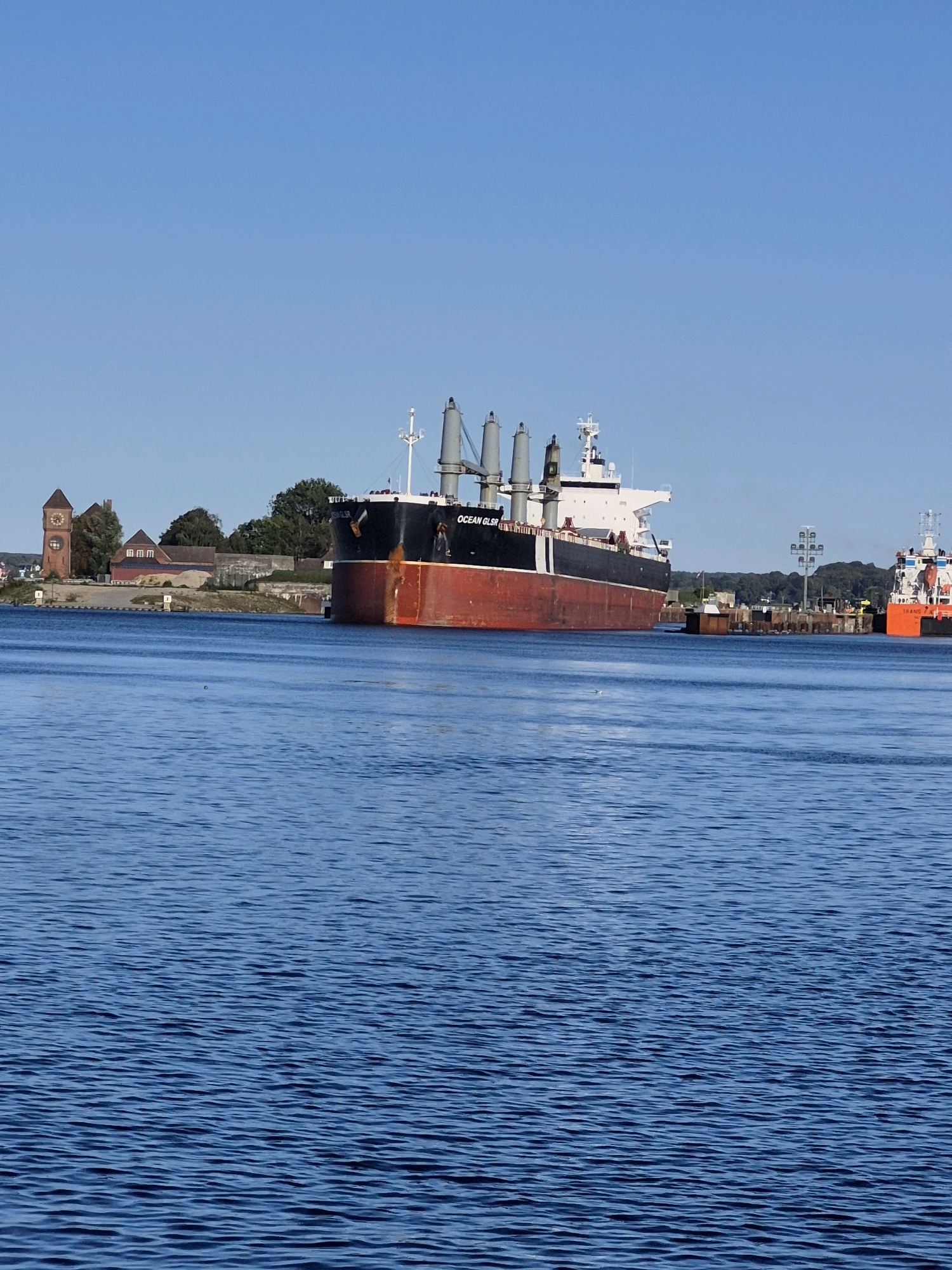 A large cargo ship, the "OCEAN GLSR", is docked in the port of Kiel, Germany. The ship has a prominent red hull and a white superstructure, with several cranes visible on its deck. In the background, a historic brick building with a clock tower stands on the shore, adjacent to other industrial buildings and trees. The ship is positioned in a calm blue body of water, likely a harbor or canal, with a clear blue sky above. Another smaller orange ship is visible in the distance on the right.