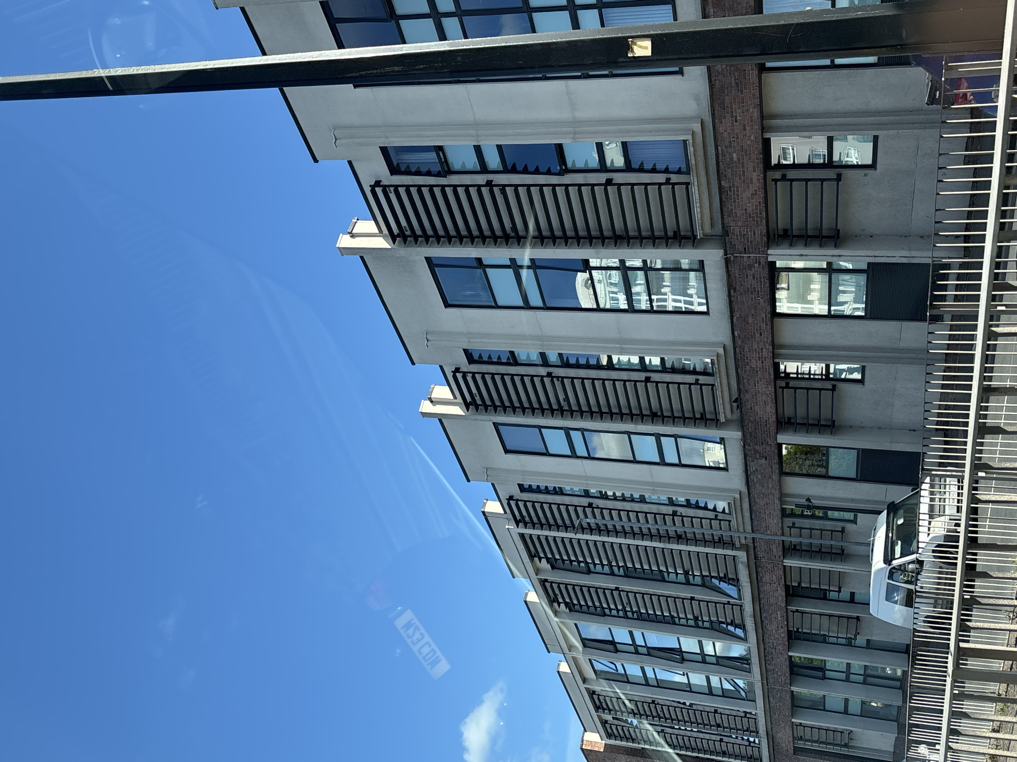 The image shows a modern building with multiple floors and rectangular windows, each featuring horizontal slats over them, taken from a low angle, looking upwards. The building's facade alternates between grey concrete and brick sections. A white van is visible at the bottom right of the frame, partially obscured by a grey metal railing. The sky above is bright blue with a few faint clouds. The image is taken from inside a vehicle, as evidenced by the reflection of a registration plate (KS3 COA) on the window. The building is a modern structure with distinct architectural design elements. This image was taken in Chester, United Kingdom.