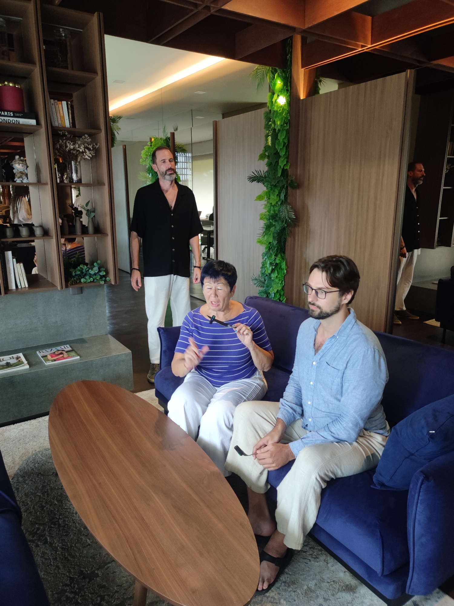 This image captures a candid moment of a discussion or interview taking place in a modern and stylish interior space, possibly a hotel lounge, office, or upscale home in North Kuta, Indonesia.
Three adults are present. In the foreground, an older woman with short dark hair, wearing a blue and white striped shirt and white pants, sits on a plush blue sofa. She is actively speaking and gesturing with her hands, with a small microphone clipped to her shirt. Beside her on the sofa, a younger man with a beard and glasses wears a light blue button-up shirt and light-colored pants. He is looking to the right, seemingly engaged in the conversation.
Standing behind the sofa is another bearded man in a black shirt and white pants, observing the interaction. His reflection is visible in a large mirror on the right side of the room, which features wooden wall panels and a vertical green plant installation.
The setting is characterized by contemporary design elements, including a rich blue sofa, a sleek oval wooden coffee table, and a wooden shelving unit filled with books and decorative items. Visible book titles include "PARIS" and "LONDON," while magazines on a low shelf display "DIOR" and "BAZAAR." Extensive green foliage, both as a vertical garden and potted plants, adds a natural, vibrant touch to the space, complementing the wooden ceiling beams and warm ambient lighting. The overall atmosphere suggests a relaxed yet professional environment.