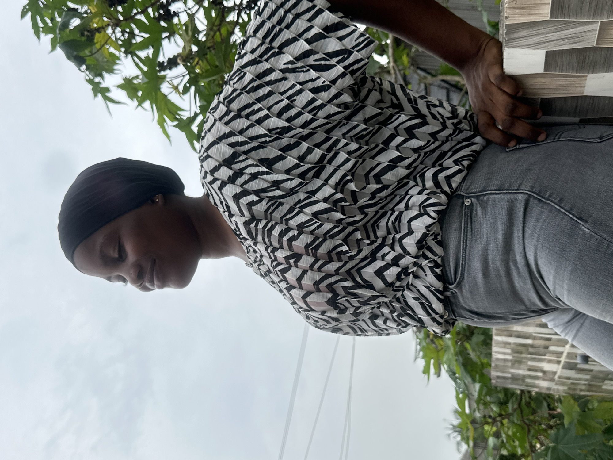 The image shows a young person, likely a woman, of dark complexion, captured in a tilted, low-angle perspective, giving a sense of looking up at her. She is wearing a black head covering (possibly a durag or headwrap), a striking black and white pleated shirt with a geometric or zebra-like pattern, and grey denim jeans.
The person is positioned against a background of an overcast sky and lush green foliage, suggesting an outdoor setting. Her head is bowed, and she is looking downwards with a calm and serene expression. A small nose stud is visible on her right nostril, and a small earring is in her right ear. Her right arm is bent, and her hand rests gently on a multi-toned grey and white tiled wall or surface, visible on the right side of the frame. The shirt's distinctive pleated texture and bold pattern are prominent, with some areas appearing slightly sheer.
The lighting indicates daytime with an overcast sky, creating soft, diffused light without harsh shadows. The overall atmosphere appears peaceful. There is no visible text in the image. The location is specified as Badagry, Nigeria.