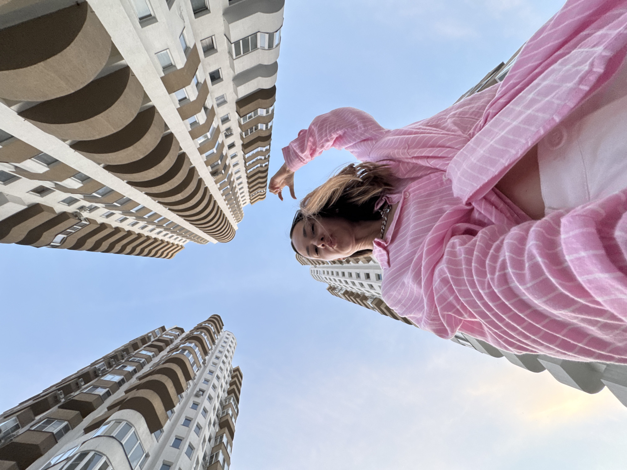 In Sochi, Russia, a young woman wearing a pink and white striped oversized button-down shirt and white pants stands outdoors, posing for a low-angle shot. She is looking directly at the camera with her lips pursed, holding her left arm up in a gesture. Behind her, two modern, multi-story buildings with curved balconies dominate the sky, set against a clear blue backdrop. The perspective emphasizes the towering architecture and the woman's presence within the urban landscape.