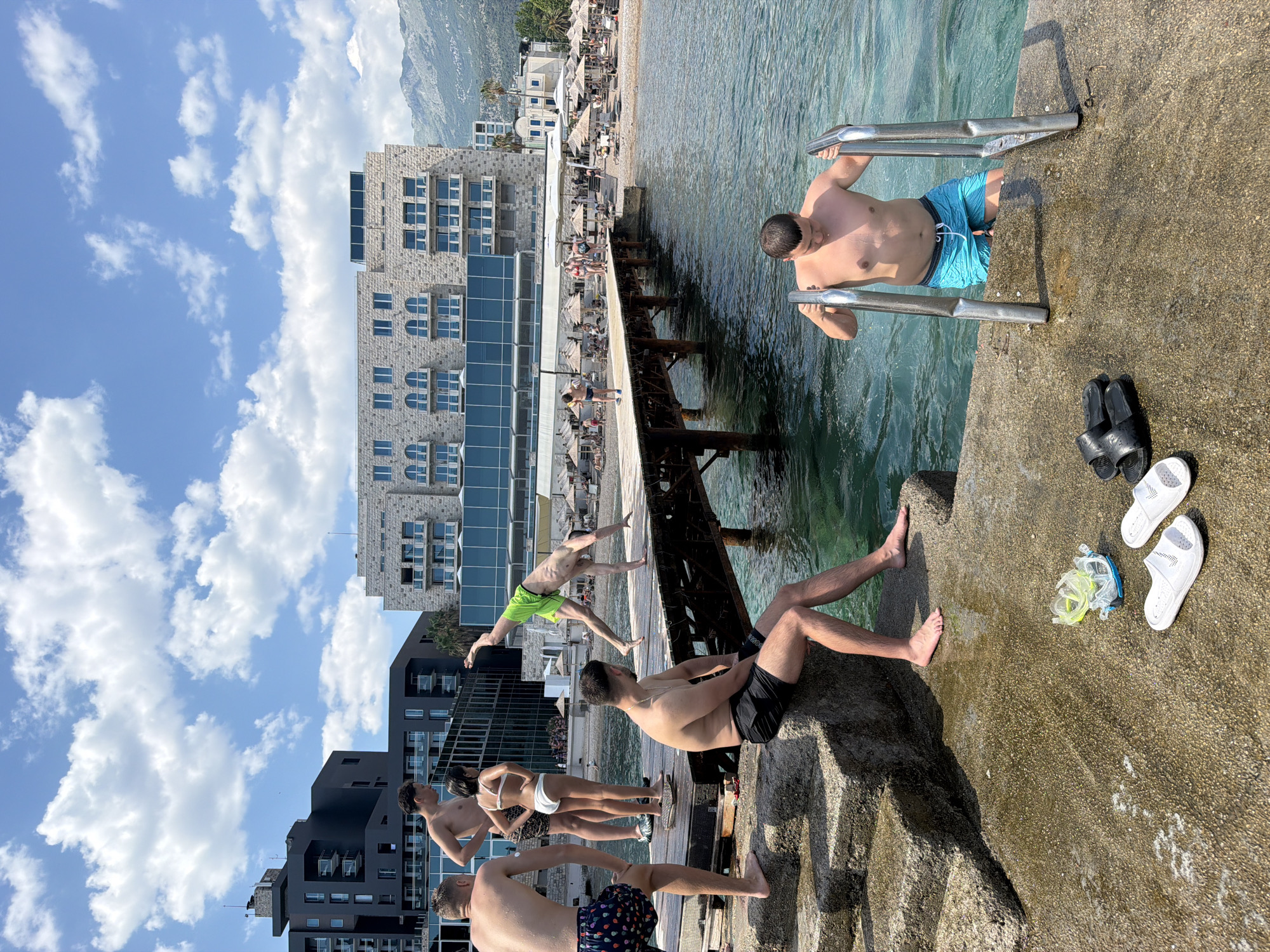 The image depicts a scene at the beach in Budva, Montenegro, with a focus on a wooden pier extending into the clear turquoise water. Several individuals are present, engaged in various activities. On the left, two men and a woman stand near stone steps leading into the water, while another man is seated on the steps. In the center, a person is performing a handstand on the pier. Further down the pier, several people are visible walking and relaxing under umbrellas on the beach. In the foreground, a man is ascending from the water using a ladder. Beach accessories, including flip-flops and snorkeling gear, are scattered on the concrete in front of the seated man. The background reveals a modern hotel building and mountainous terrain. The sky is partly cloudy.
