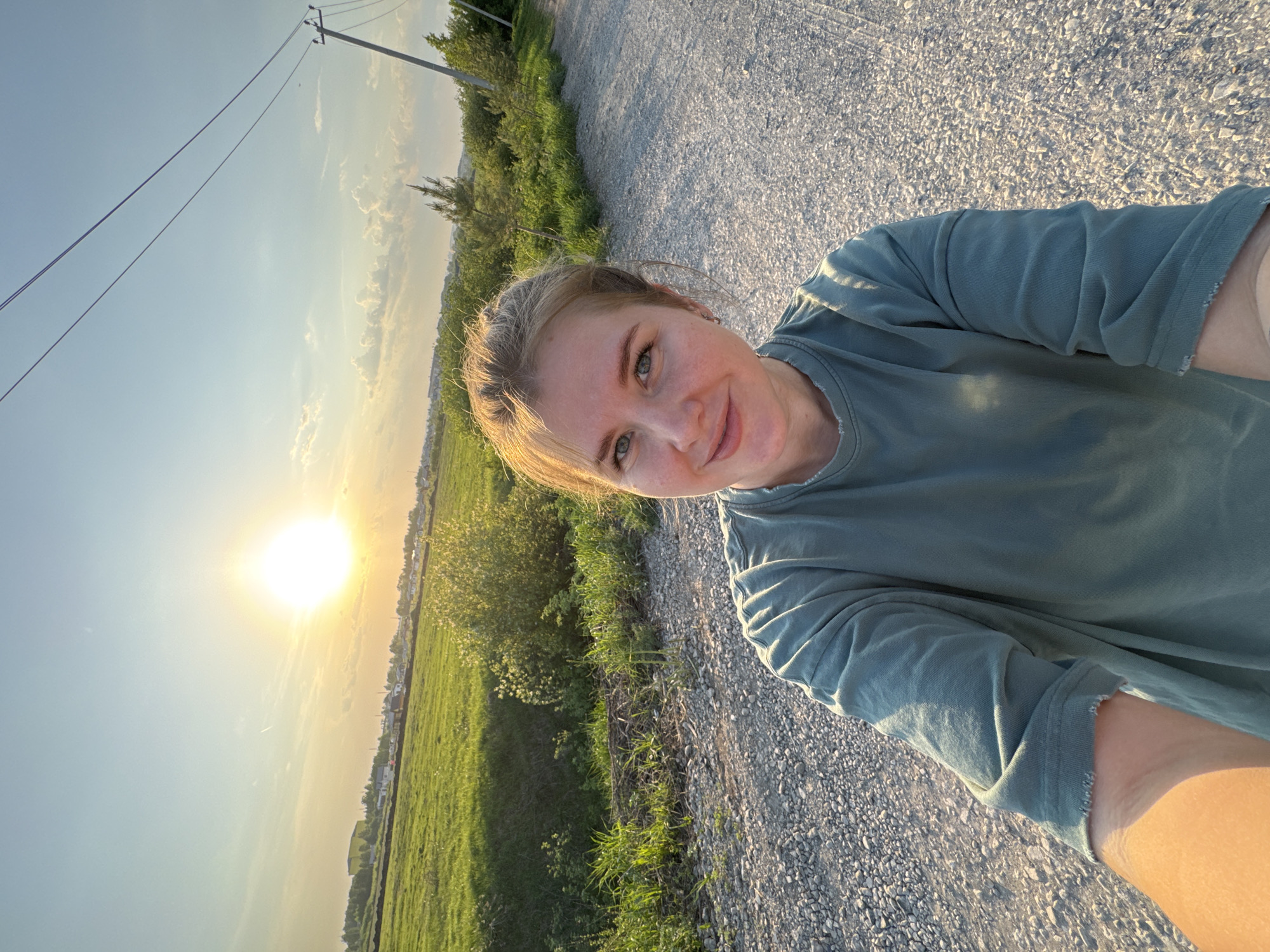 A selfie taken outdoors features a fair-skinned woman with blonde hair wearing a teal t-shirt, standing on a gravel road. The sun is positioned low in the sky, suggesting either sunrise or sunset. A field is visible to the left of the road, and vegetation lines the roadside. A power pole stands in the background on the left side. The ground consists of compacted gravel. The location is assumed to be Горный, Russia, based on provided information.
