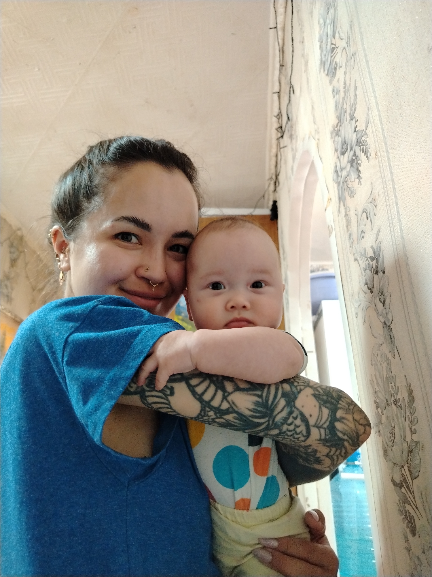 A woman is holding a baby. The woman has her hair pulled back, a nose ring, earrings, and a tattoo on her arm. She is wearing a blue shirt. The baby is wearing a patterned shirt and yellow pants. They are indoors in a room with floral wallpaper and a string of lights. The location is in the city of Unknown, Unknown.