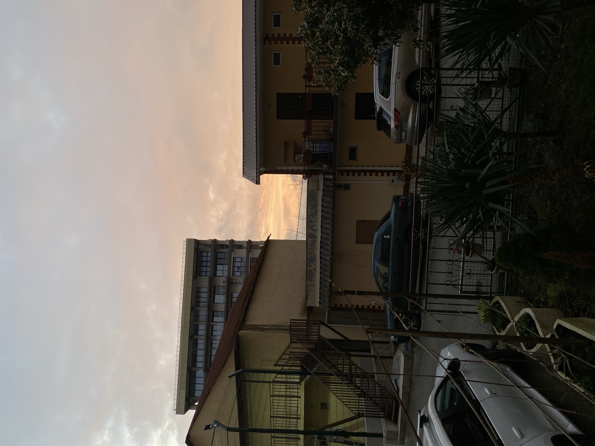 The image depicts an urban scene, likely at dawn or dusk, given the soft, pastel sky. The setting appears to be in Sochi, Russia, due to the architectural style and the presence of subtropical vegetation.
Two buildings are prominent. One is a two-story building with a yellow facade and a metal roof. It has small balconies with metal railings and what appears to be flower boxes. The second building is taller, of concrete construction, with multiple stories and numerous windows, possibly an apartment or office building. A rusty metal staircase is visible against the side of this building.
Between the buildings, a strip of orange-colored sky is visible.
There are two cars parked on the side of the road: one grey SUV and one white sedan. The SUV is parked adjacent to the yellow building.
The area in front of the buildings is fenced. Tall palm-like plants and other subtropical greenery are visible within the fenced area.
No people are visible in the image.