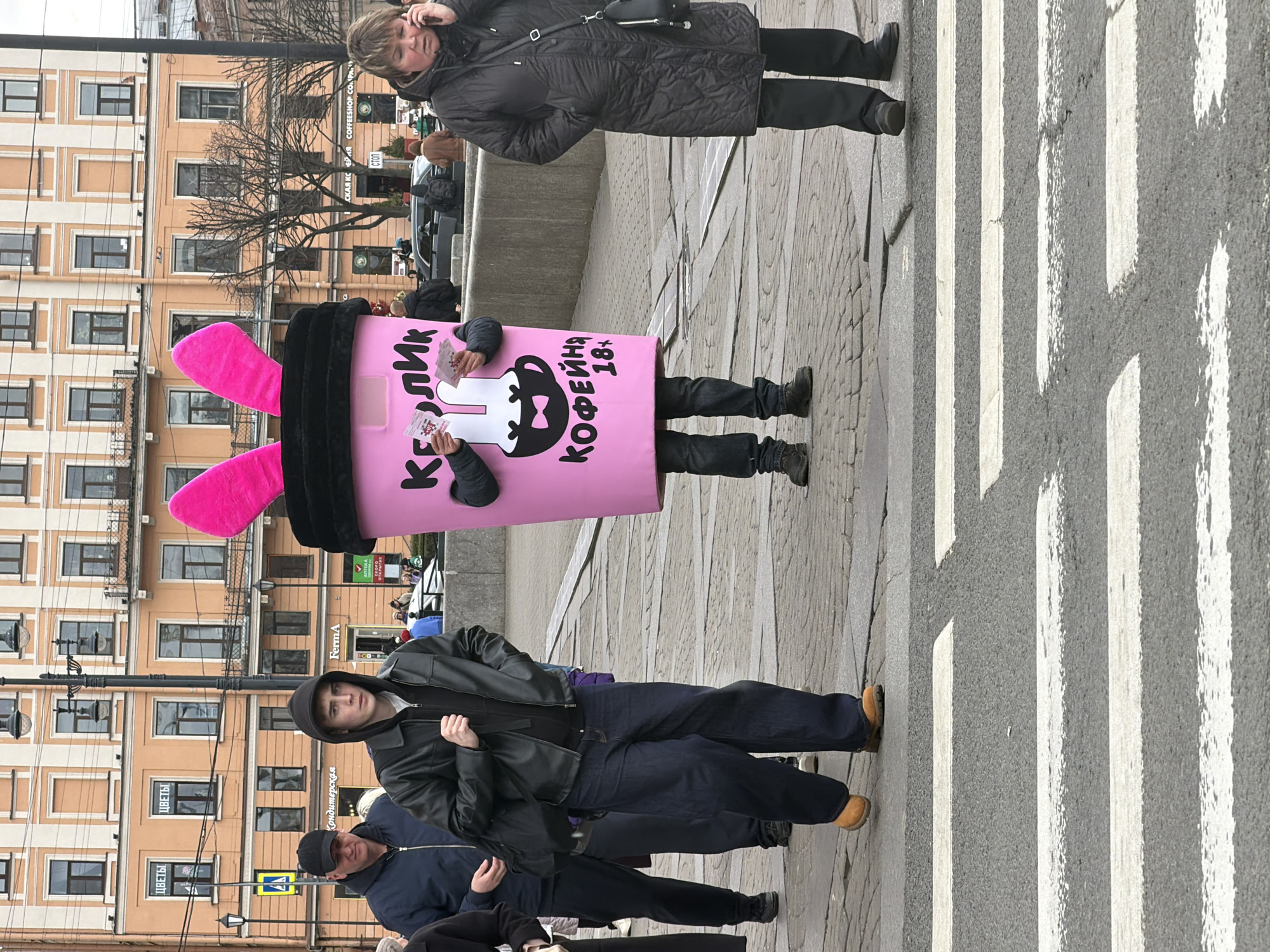 This full-length photograph captures a vibrant street scene in Saint Petersburg, Russia, likely during cool weather as indicated by the bare trees and heavy coats worn by pedestrians. The image is taken from a slightly low angle, showing a paved sidewalk and a zebra crossing on the right.
The central figure is a person dressed in a large, eye-catching pink coffee cup costume, adorned with black trim at the top and two prominent pink bunny ears. The costume features a cartoon rabbit design and Cyrillic text that reads "КЕЛИК КОФЕЙНЯ 18+" (Kelik Coffee Shop 18+), suggesting it's a promotional mascot for a coffee shop. This individual is holding small white cards or flyers, presumably for distribution.
To the right of the costume, a woman in a black quilted coat and dark pants is seen talking on a cell phone, carrying a black shoulder bag. In the foreground on the left, two men are visible; one in a black leather jacket over a hoodie looks directly at the camera, while another in a dark blue jacket and black baseball cap is partially seen.
The background features several multi-story buildings with light-colored facades and numerous windows, characteristic of European urban architecture. Visible signs on these buildings include "ЦВЕТЫ" (Flowers), "Кондитерская" (Confectionery), "FermA", and fragmented text like "COFFEESHOP CO" and "ОТКРЫТИЕ" (Opening), indicating commercial establishments. A "СТОП" (STOP) sign is also present. The sky appears overcast, suggesting daytime with soft, diffused light. The scene depicts a bustling urban environment with individuals engaged in daily activities.