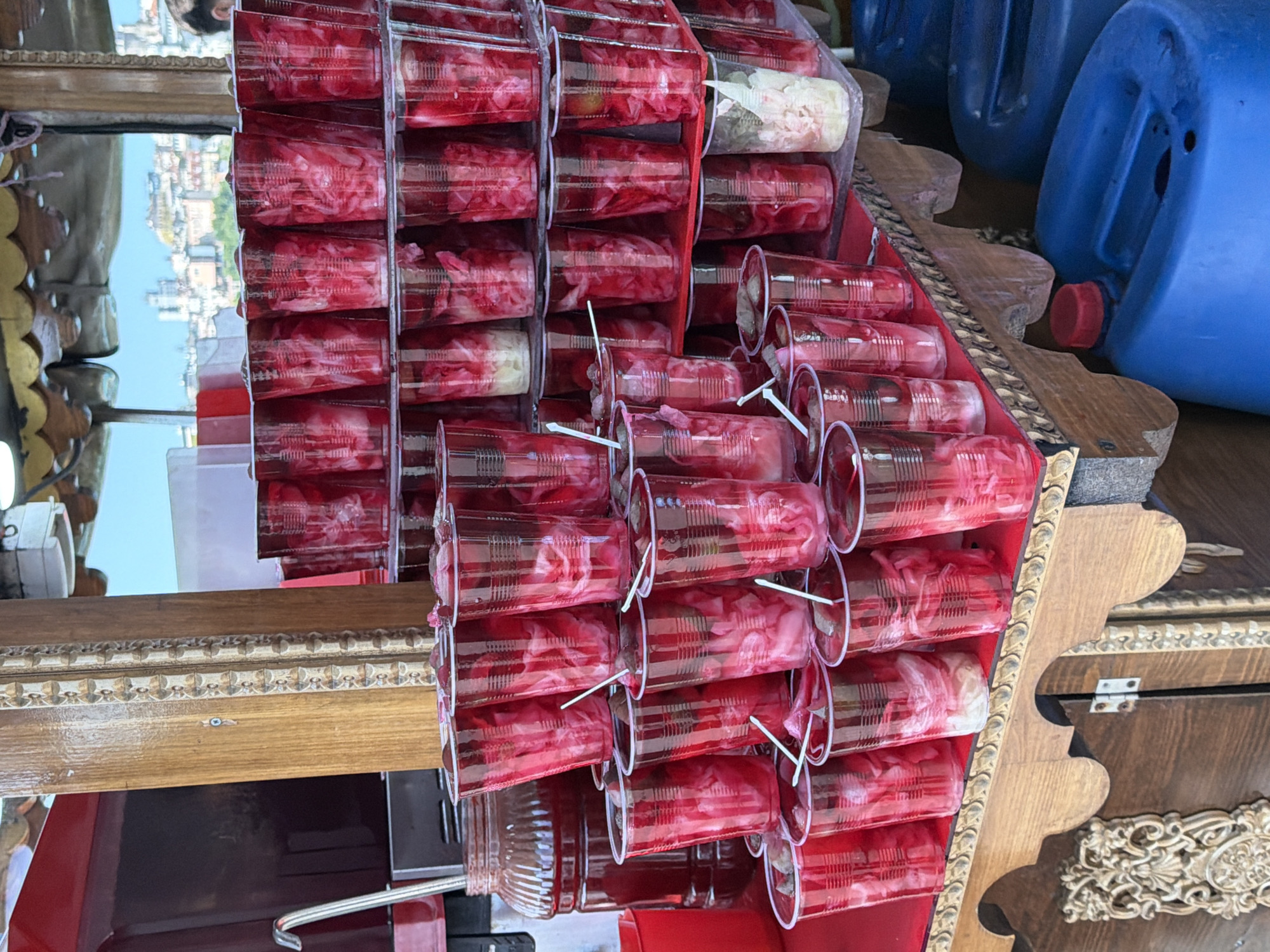 The image shows a street food stall in Fatih, Istanbul, Turkey. The stall is displaying many plastic cups filled with a pinkish-red liquid and shredded vegetables, likely pickled turnips, a common Turkish street food. The cups are arranged on wooden shelves. Behind the cups, a large glass jar filled with the same red liquid is visible. A red ladle rests on the edge of the jar. The stall itself has a rustic wooden structure with some decorative carvings. In the background, a cityscape with buildings and a bridge can be seen under a bright blue sky, suggesting an outdoor location. Blue plastic containers are stacked to the right of the stall.