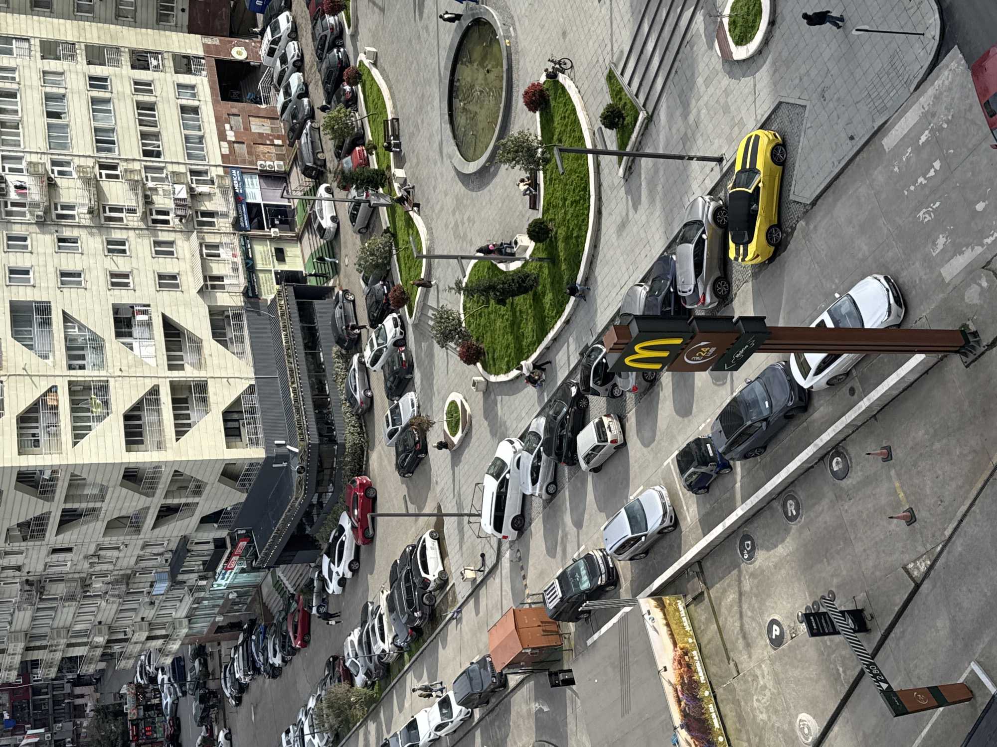 The image displays an elevated view of a bustling urban street and plaza in the city of Unknown, Unknown, under daylight conditions. Multi-story commercial and residential buildings form the backdrop, with a light-colored structure on the left featuring balconies and ground-level businesses, including "AMERICAN MEDICAL CENTER" and a "RESTAURANT PRIME TIME" sign in a non-Latin script. An adjacent building exhibits a distinct facade with angular geometric patterns.
A large paved public plaza occupies the mid-right, defined by crescent-shaped raised garden beds planted with green grass, small trees, and reddish shrubs. A circular, shallow fountain containing stagnant water is situated in the upper right section of the plaza. Multiple dark-colored benches are positioned along the green spaces.
The foreground and left side feature a commercial area centered around a McDonald's establishment, identified by a tall brown pole with a yellow "M" logo and a "24h McDrive" sign. A drive-thru lane is visible, along with a gated parking entrance marked "SOCAR" in the lower left.
Numerous vehicles, including sedans, SUVs, and vans, are parked densely along the street, in designated bays, and within parking lots throughout the scene. A yellow Chevrolet Camaro convertible with black racing stripes is prominently parked near the McDonald's sign. An electric scooter is parked on a sidewalk, and a bicycle is visible near steps leading into the plaza.
Approximately 20-30 pedestrians are dispersed across the scene, walking on sidewalks, navigating the plaza, or seated on benches. No distinct interactions between individuals are discernible.