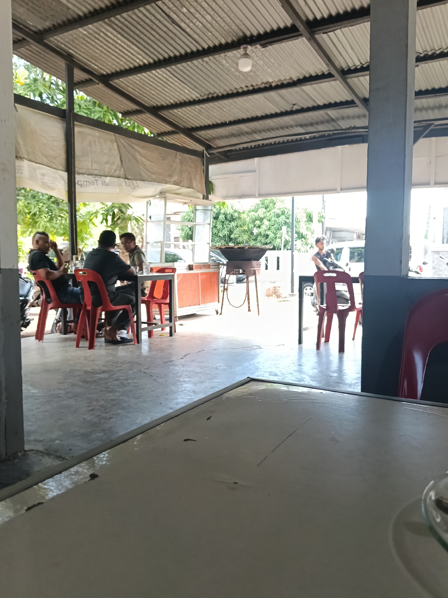 The image depicts a casual outdoor dining area, likely a warung or a roadside eatery, in Banda Aceh, Indonesia. Three men are seated at a table with red chairs, engaged in conversation. The setting is open-air, covered by a corrugated metal roof supported by a pillar. Tropical foliage is visible in the background, suggesting a warm climate. One man on the far left wears a black t-shirt and jeans. The man in the center wears a dark t-shirt and dark pants. The third man is partially obscured, wearing a light-colored shirt. A wok and stand are visible further back, hinting at food preparation. The ground is paved concrete. The overall impression is one of relaxed daily life in a common Indonesian urban or semi-urban setting.