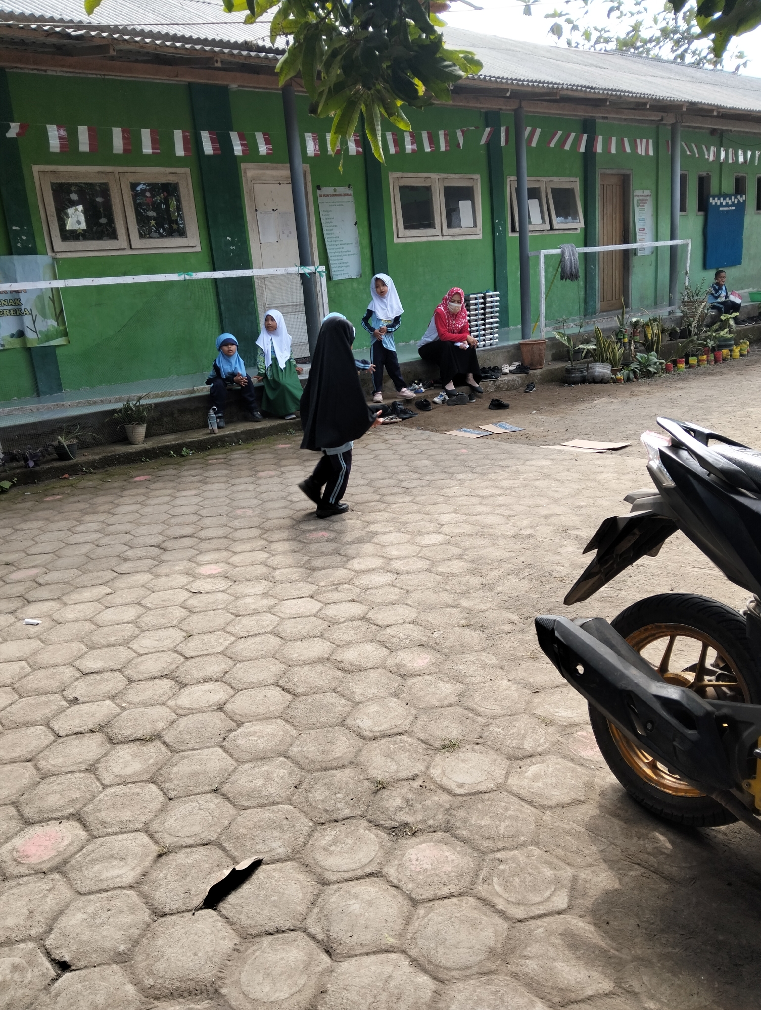 The image depicts an outdoor scene in front of a green building with a corrugated metal roof, likely in Desa Wringin Putih, Indonesia. A group of young people, mostly wearing hijabs, are present. Two girls in hijabs are seated on steps in front of the building, one in a blue hijab and the other in green. Another person wearing a red hijab and a face mask is also seated. A person in a black hijab and black pants is walking away from the camera. The building's facade features windows and a door, with red and white striped flags decorating the upper portion. The ground is paved with hexagonal concrete blocks. In the foreground, the rear of a black motorcycle is visible, showing a wheel with a gold rim and part of the exhaust system. The environment appears to be a school or community building with some potted plants and scattered items on the ground, including shoes and cardboard. The lighting suggests it is daytime.