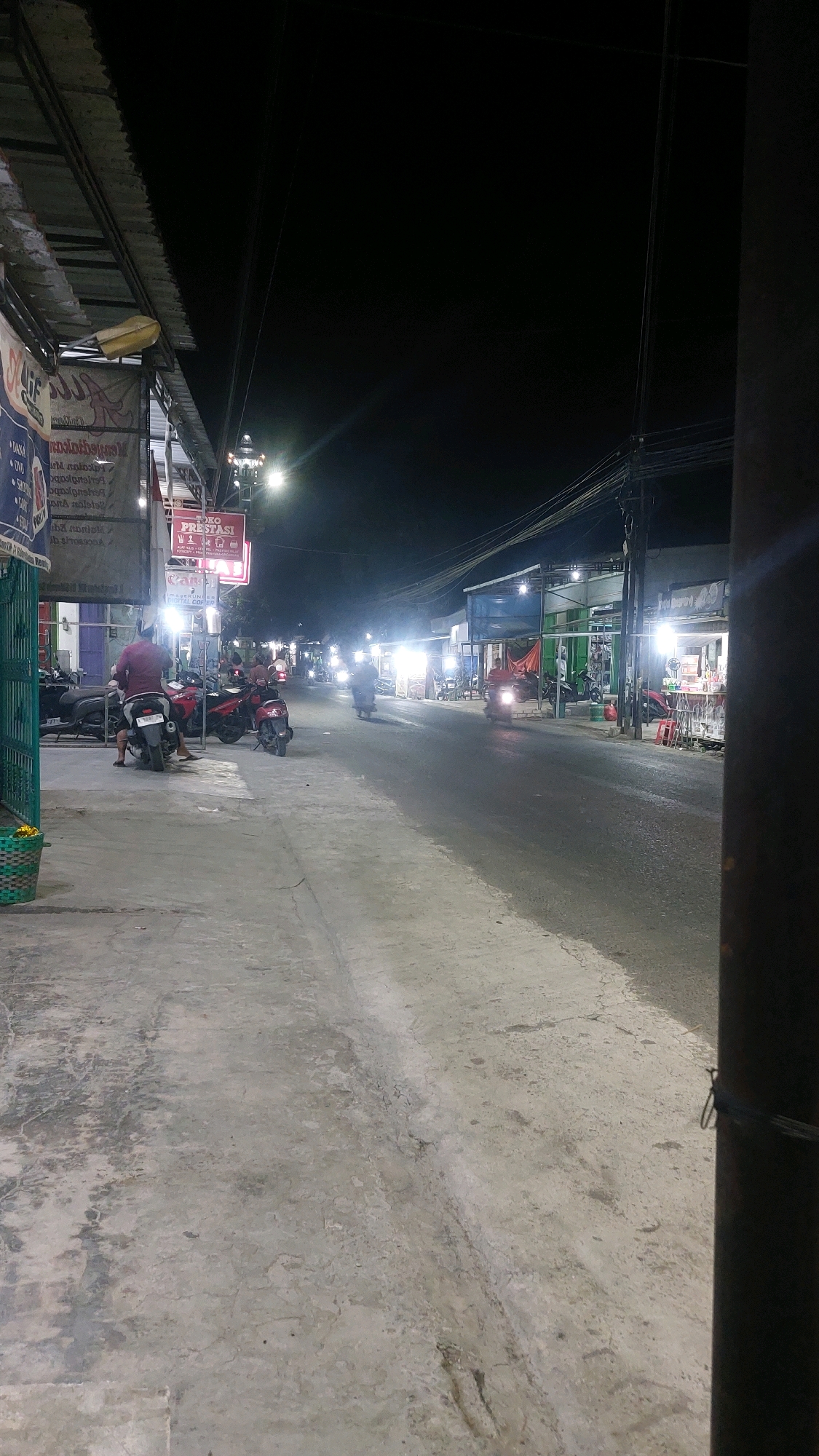 The image depicts a street scene at night in Karas, Indonesia. The foreground is dominated by a concrete sidewalk with some debris and cracks. To the left, a green metal fence encloses a small area with a woven basket. A person wearing a maroon shirt is seated on a motorcycle, which is parked in front of a row of shops. Several other motorcycles are also visible, some parked and some in motion on the street. The shops lining the street are illuminated by bright lights, displaying signs and merchandise. Traffic, consisting of motorcycles, is moving along the asphalt road. Power lines and poles are visible crisscrossing the sky, indicating an urban or semi-urban environment. The overall atmosphere is that of a bustling commercial area operating late into the evening.