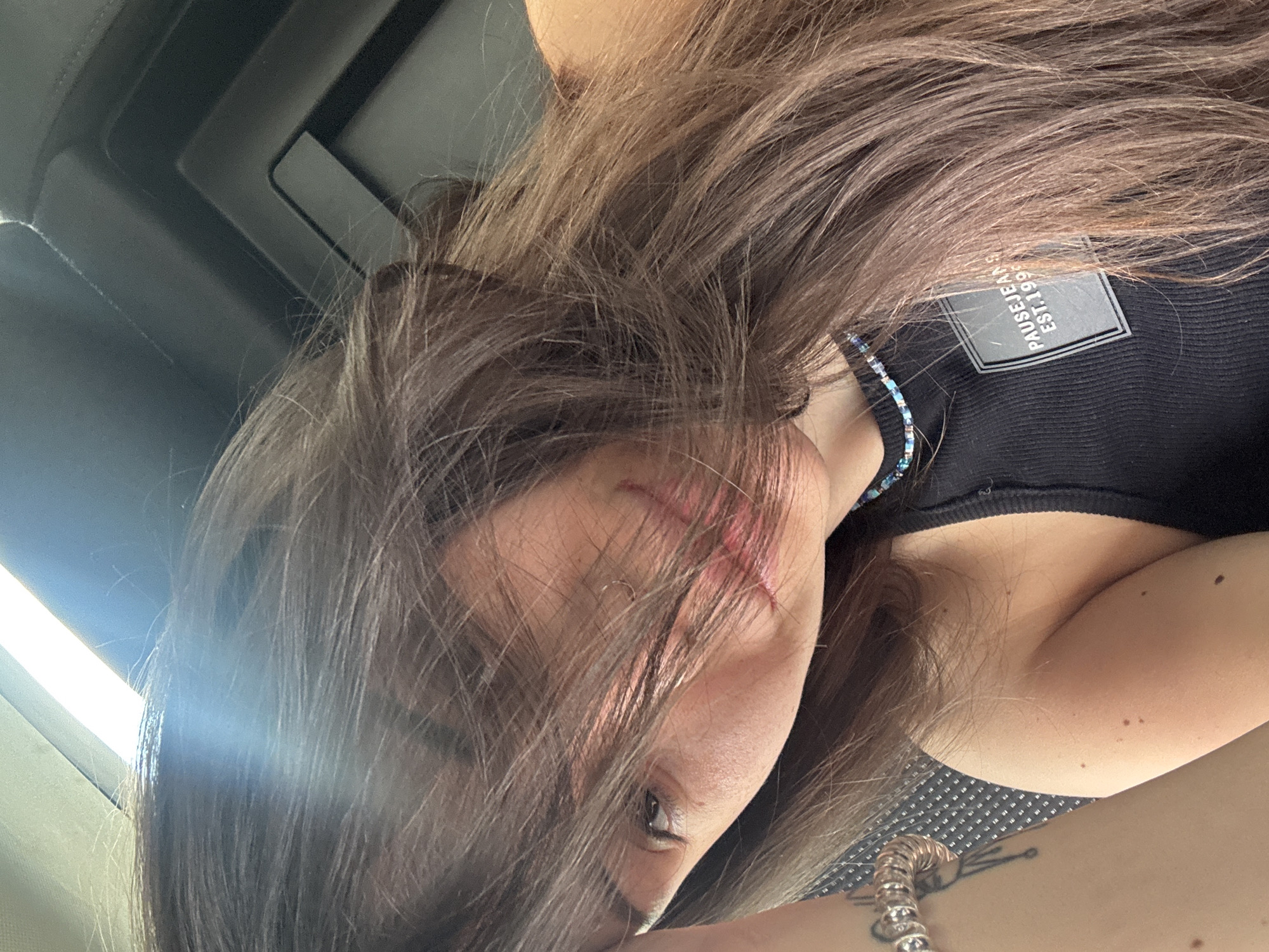 The image, rotated approximately 90 degrees counter-clockwise, presents a close-up of a person, likely female, captured within the interior of what appears to be a vehicle. Her long, brown hair, streaked with lighter tones, cascades across her face, partially obscuring it. One eye is visible, looking upwards towards a bright light source. A reddish, abrasion-like mark is noticeable near the left corner of her mouth, partly hidden by her hair.
She is wearing a black ribbed tank top or camisole. A small, delicate necklace made of blue and white beads is visible around her neck. On her top, a rectangular label or patch clearly displays the text: "PAUSE JEANS EST. 1999".
In the lower right portion of the frame, part of her arm is visible, adorned with a metallic bracelet, possibly of a coiled design, and a stylized tattoo that resembles an abstract shape or insect. The background in the upper left reveals the dark, structured ceiling of a car or bus interior. A very bright light, likely sunlight streaming through a window, creates a prominent lens flare on the left side, indicating it is daytime. The pattern on the seat upholstery in the lower right further suggests a vehicle setting. The overall feel is that of a casual, candid snapshot. While the location context provided is Sofia, Bulgaria, the image itself does not contain specific visual cues to confirm this geographical detail. The person's expression is somewhat obscured by her hair, but she appears relaxed, looking towards the bright light.