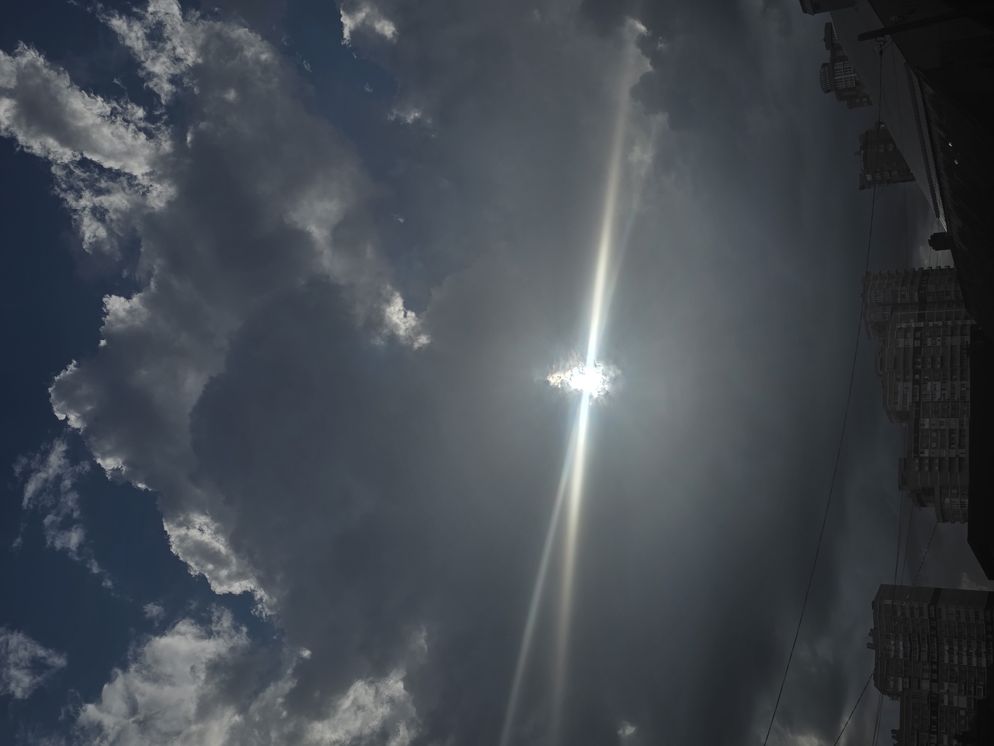 The image presents a sky filled with clouds, ranging from dark, ominous storm clouds to lighter, more scattered ones. The sun is partially obscured by the clouds, creating a bright flare and visible sunbeams. On the right side of the frame, there are buildings, suggesting an urban environment. Given the city of Krasnodar, Russia is specified, it's inferred the photograph was taken in Krasnodar. The perspective is looking upwards.