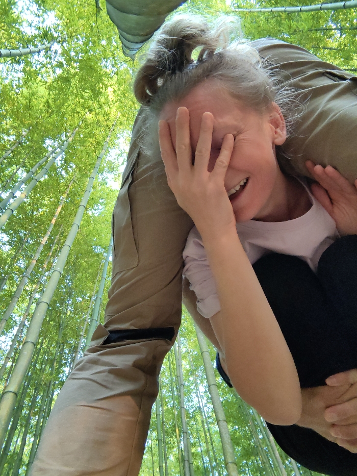 An adult female is depicted being carried upside down over the left shoulder of another individual in a bamboo forest in Mù Cang Chải, Vietnam. The female, with light-colored, disheveled hair, is positioned with her head towards the lower right of the frame. She is visibly laughing, her mouth open and eyes squinted, and her right hand is covering the lower portion of her face. She wears a light-colored short-sleeved top and dark-colored pants. Her left hand is visible, gripping the carrier's leg. The carrier's left leg, clad in light brown cargo-style pants with a dark horizontal strap, is prominent in the center-left. The background is filled with numerous tall, slender green bamboo stalks, extending vertically towards a bright sky visible through the canopy.
