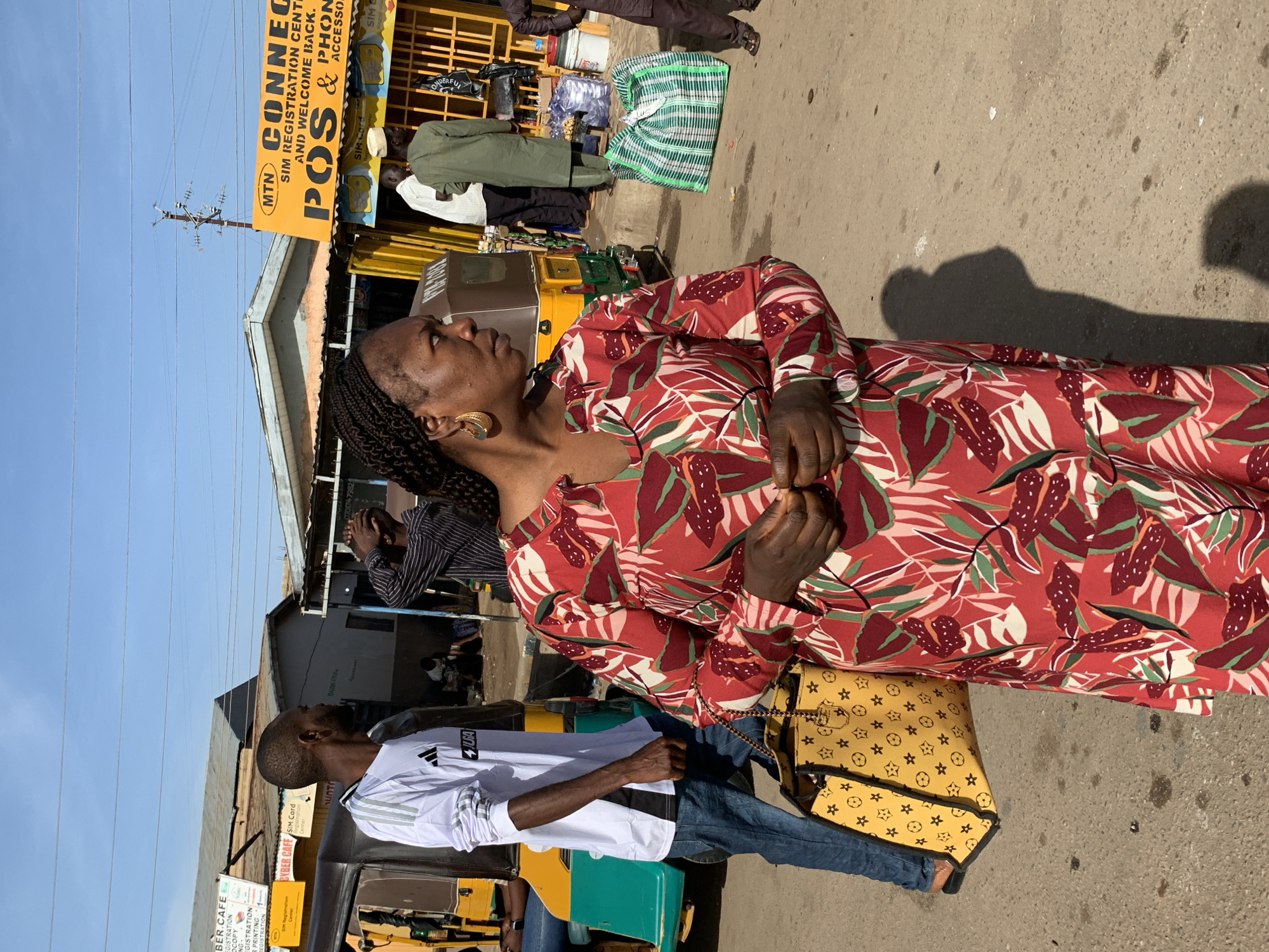 This image captures a vibrant street scene in Gura Top, Nigeria, under bright, clear daylight, suggested by the blue sky and distinct shadows on the ground. The setting appears to be a bustling market or commercial area with various stalls and small businesses.
In the foreground, a woman with dark skin and braided hair is prominent, wearing a red, green, and white tropical-patterned long-sleeve dress and large gold hoop earrings. She carries a yellow shoulder bag adorned with star and diamond patterns. Her gaze is directed upwards and to the left, away from the camera. Beside her, a man with short dark hair is partially visible, wearing a white long-sleeve shirt with black stripes and blue jeans, standing next to a yellow and green three-wheeled auto-rickshaw, commonly known as a tuk-tuk.
The background features several market stalls or small shops. One stall has an olive-green garment displayed. Another figure in the background has their hands near their face, possibly shielding from the sun or engaged in an activity. Various goods, including plastic bags and other items, are visible near the stalls.
Visible text on signs indicates commercial activities:
* A large yellow sign advertises "MTN CONNECT SIM REGISTRATION CENT AND WELCOME BACK. POS & PHONE ACCESSO."
* Another sign for a "CYBER CAFE" offers "SIM Card Registration."
* Further text includes "BER.CAFE REGISTRATION COPY PRINTING REGISTRATION," "DAGES TRIPA," "JUGA," and a phone number "0701747982."
* A bag on a stall has the word "WONDERFUL" printed on it.
The overall scene depicts everyday life in a lively urban or semi-urban market environment, with people moving about and engaging in commerce.
