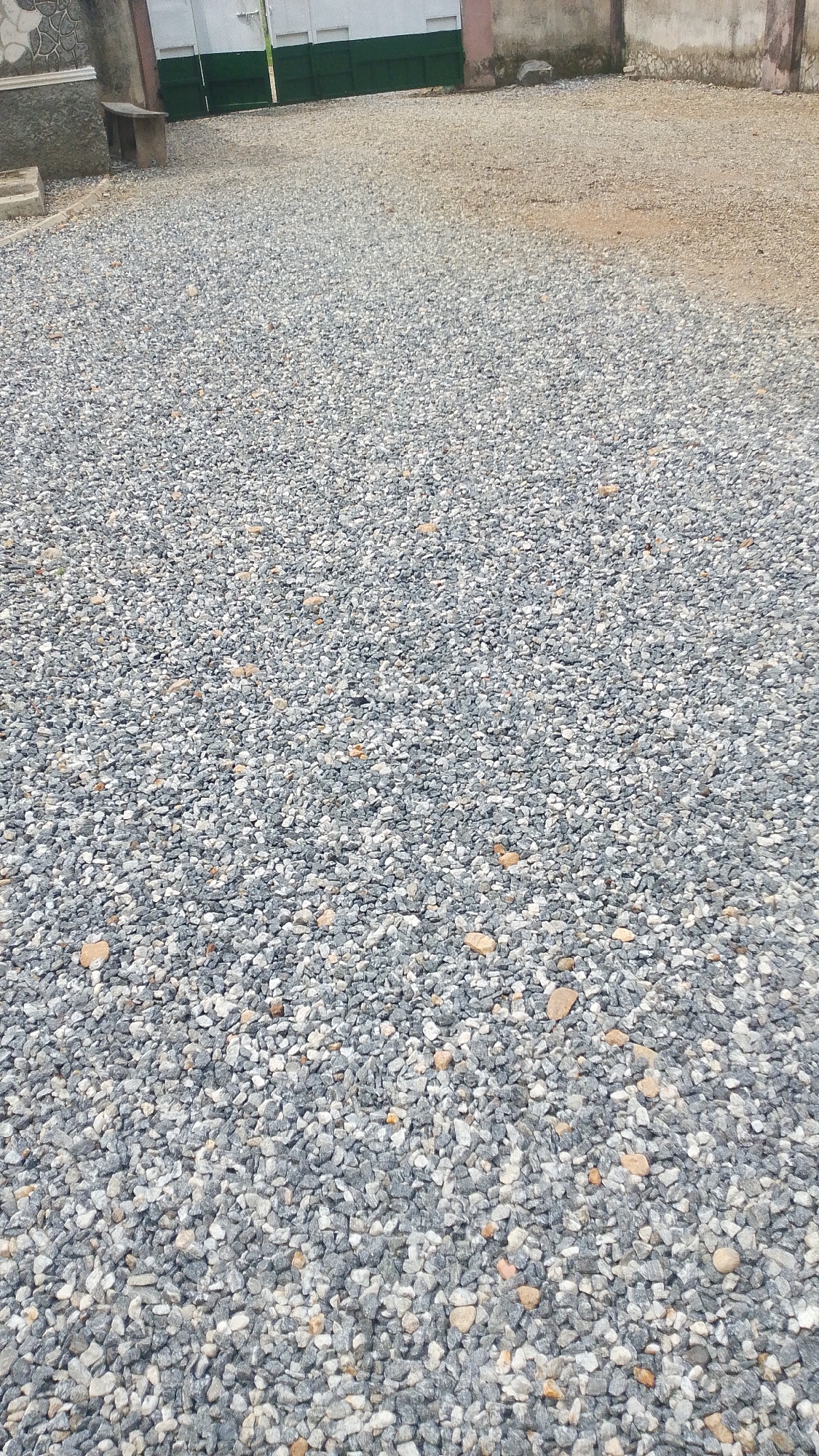 The image shows a gravel-covered ground in Balewa, Nigeria. In the background, there is a building with a green and white gate, and a concrete wall. The ground is covered with small, grey and white stones. There are no people or animals visible in the image. The scene appears to be an outdoor area, possibly a courtyard or a driveway. The lighting suggests it is daytime.