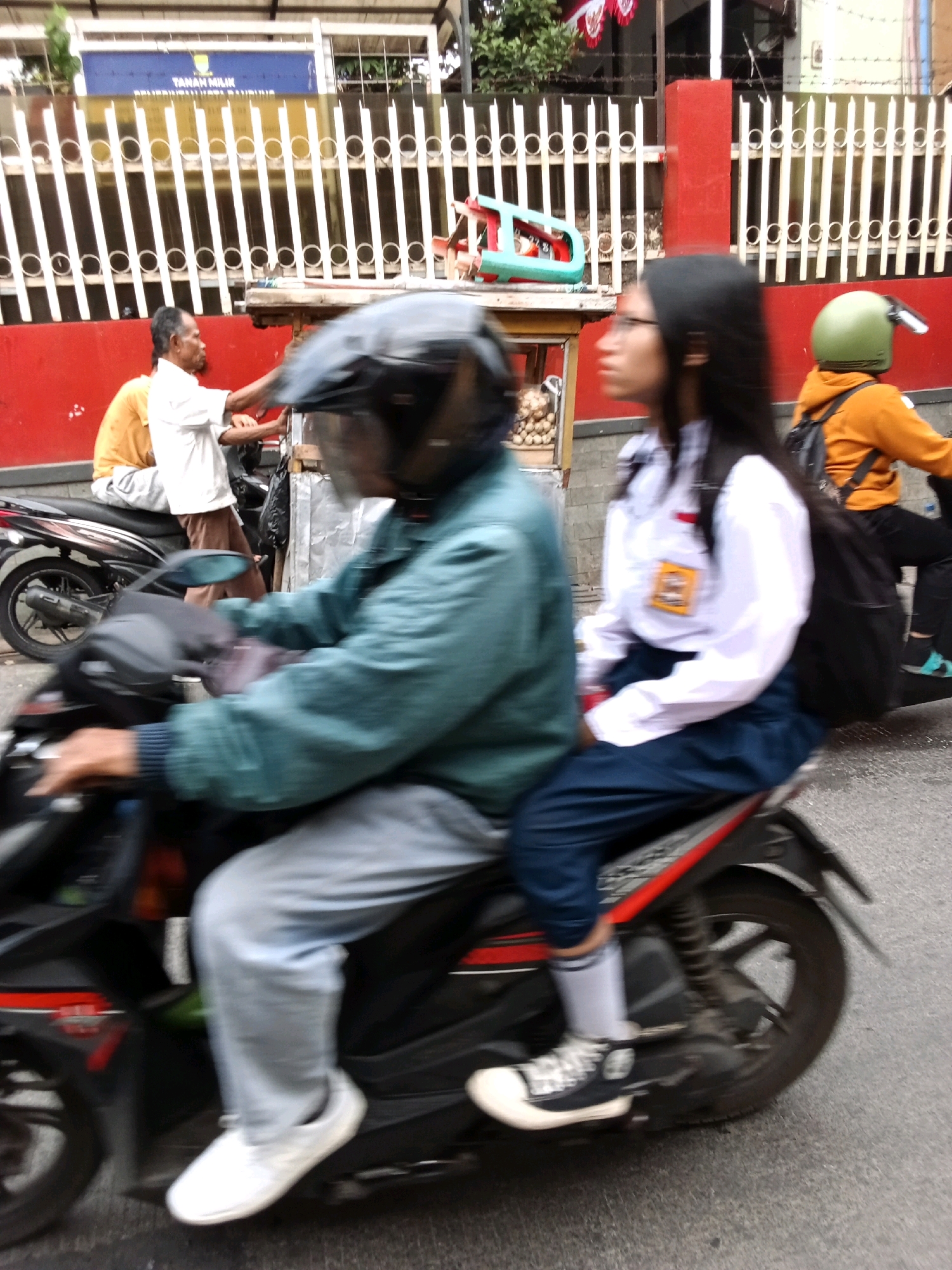The image depicts a street scene in Bandung City, Indonesia, with a focus on people riding motorcycles. In the foreground, two individuals are on a motorcycle. The person in the front is wearing a teal jacket and a black helmet with a visor. Their hands are on the handlebars, and they are wearing grey pants and white sneakers. The person behind them is wearing a white school uniform shirt with a red and yellow emblem on the chest, dark blue pants, and black and white Converse sneakers with white socks. They are carrying a black backpack.
Behind them, another motorcycle is visible, carrying two people. The rider in front is wearing an orange hooded jacket and a green helmet. They are wearing black pants and dark sneakers. The passenger behind them is wearing a yellow t-shirt and appears to be an adult male.
The background features a red wall and a white metal fence with circular motifs. A street cart with items on display is also partially visible. The road surface is asphalt. The overall impression is a snapshot of daily life in a busy urban environment in Bandung.