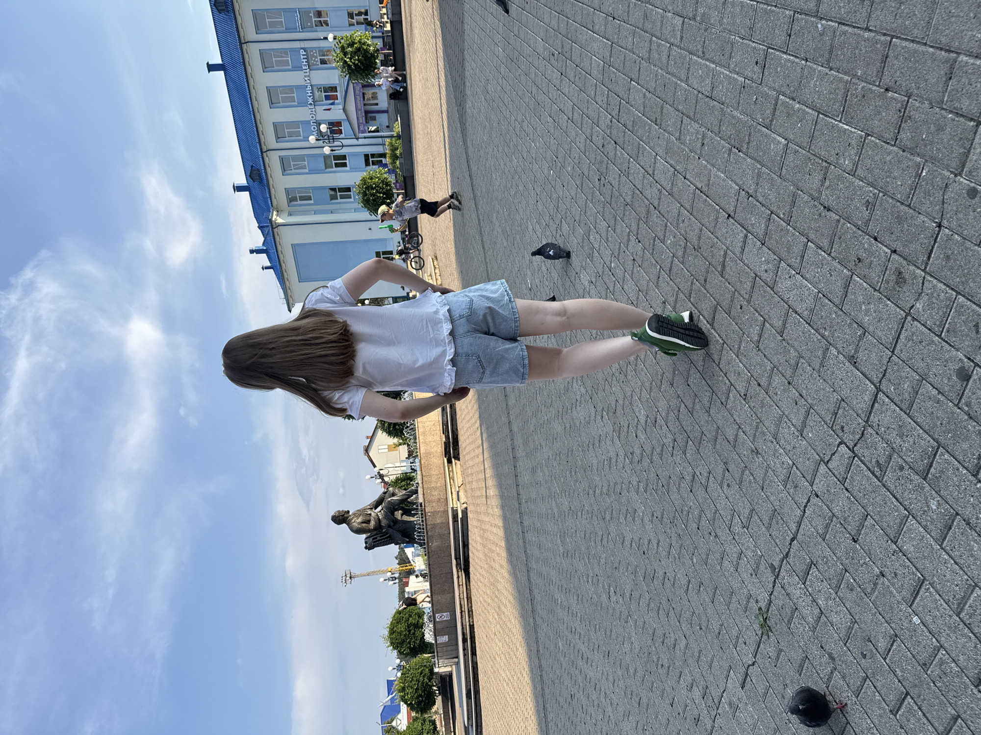 In an outdoor urban square paved with gray rectangular bricks, a young girl with long brown hair is viewed from behind, walking away from the camera. She is wearing a white t-shirt and denim shorts. To her right, a pigeon walks on the pavement. In the mid-ground, a bronze statue of a seated man is in front of a circular fountain. Beyond the fountain, a light blue building with a blue roof has the Cyrillic text "МОЛОДЕЖНЫЙ ЦЕНТР" above its entrance. A young boy in a yellow cap is running in the same direction as the girl, further to the right, holding a green object. In the background, trees and a lamppost are visible. The sky is blue with scattered white clouds.