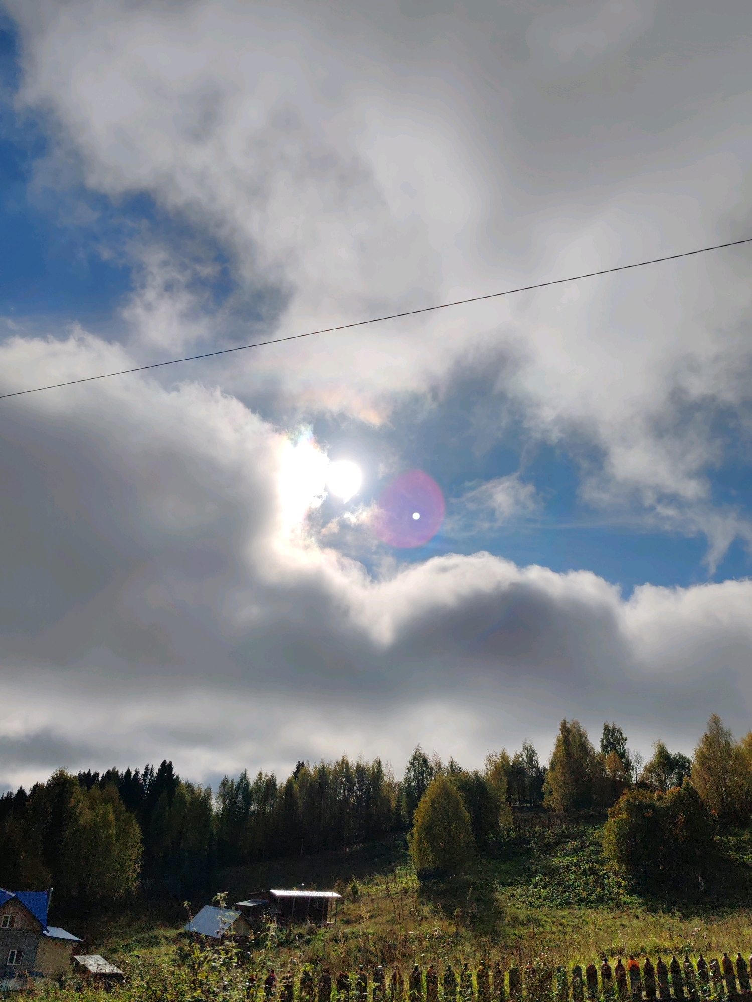 The image depicts a rural landscape, likely in or near Чухлэм, Russia, under a partly cloudy sky. The sun is visible, partially obscured by clouds, creating a bright lens flare effect and a subtle rainbow halo. A thin black wire runs horizontally across the upper portion of the sky. The middle ground features a dense line of trees, transitioning from dark green to autumnal yellows and oranges, suggesting the season is likely autumn. The foreground shows a grassy hillside dotted with small structures. To the left, a house with a blue roof and white trim is partially visible. Further to the right, a few smaller outbuildings with corrugated roofs are present. A wooden fence, appearing rustic and possibly made of posts, runs across the very bottom of the frame. The ground is covered in green grass and vegetation, with some patches of brown and yellow, consistent with autumn. No people are visible in the image. The overall scene conveys a sense of peaceful, rural life.