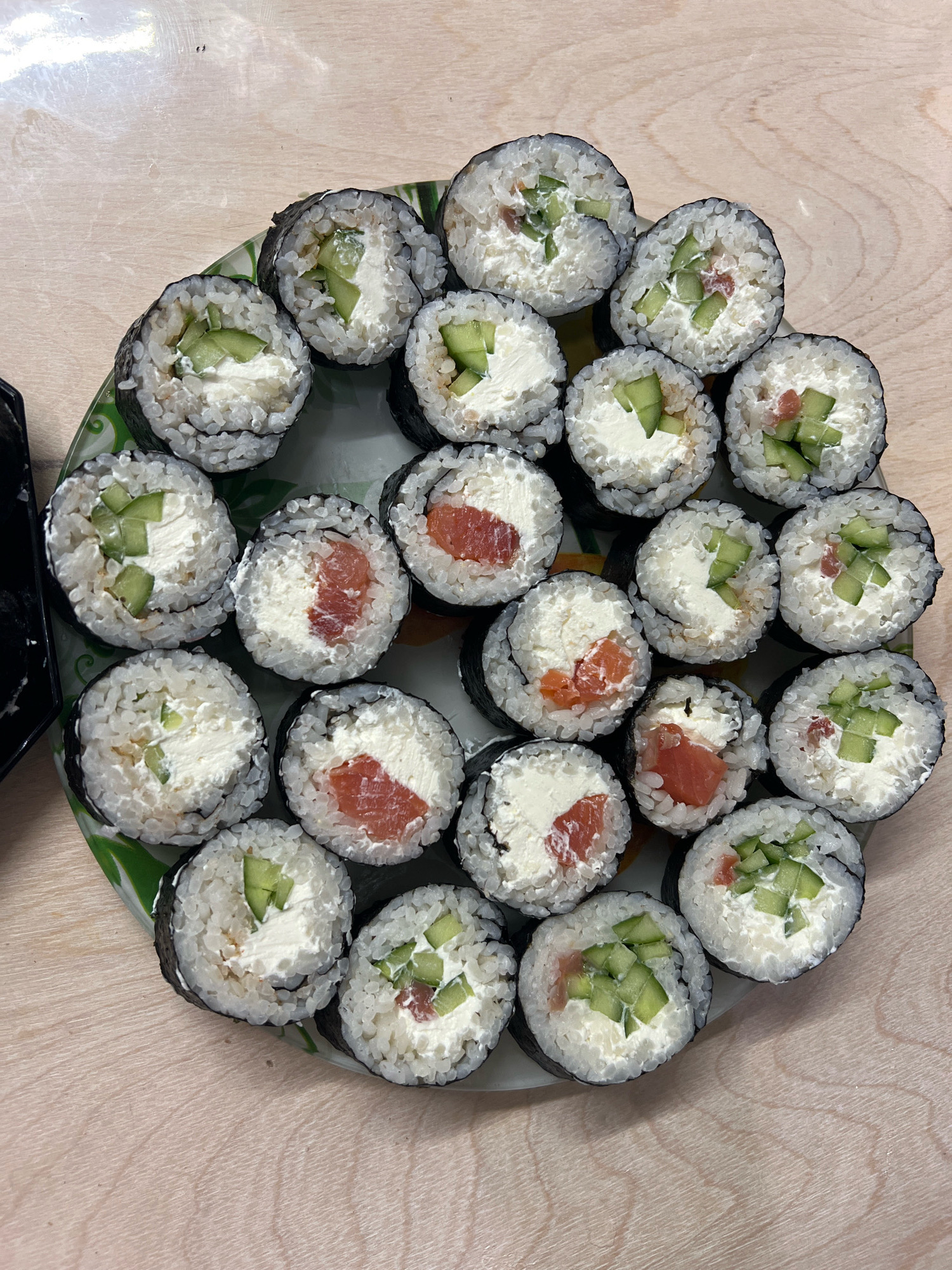 A circular arrangement of sushi rolls is displayed on a patterned green and white plate. The sushi rolls are of the maki type, characterized by their cylindrical shape with rice and fillings wrapped in seaweed. The fillings appear to be a white creamy cheese, diced cucumber, and pieces of pink salmon. The plate rests on a light-colored wooden surface with visible grain. The lighting suggests an indoor environment, likely a table setting. There are no people or other objects of significance in the immediate vicinity of the sushi.