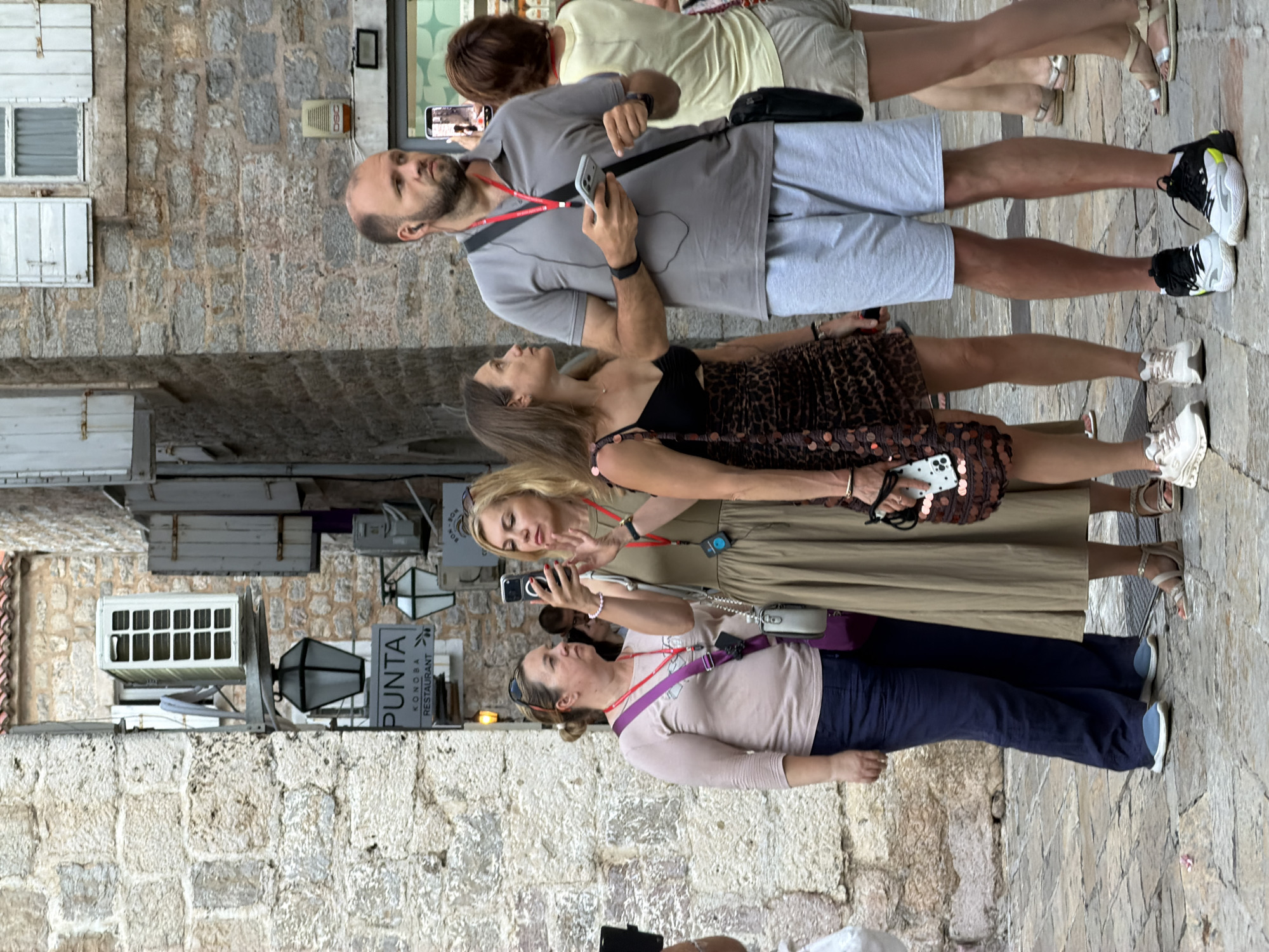The image captures a street scene in Budva, Montenegro, likely in the Old Town given the stone architecture. In the foreground, a man with a beard and earpiece, wearing a grey polo shirt and light grey shorts, holds a smartphone and looks off to his right. To his left, a woman with dark hair and a black top with a leopard-print skirt stands facing him. Behind her, a blonde woman in a tan dress is taking a photo with her smartphone. To the far left, a woman in a light pink shirt and dark blue pants is looking towards the blonde woman. The background features stone buildings with shuttered windows and a sign that reads "PUNTA KONABA RESTAURANT". The ground is paved with cobblestones. The lighting suggests it is daytime.