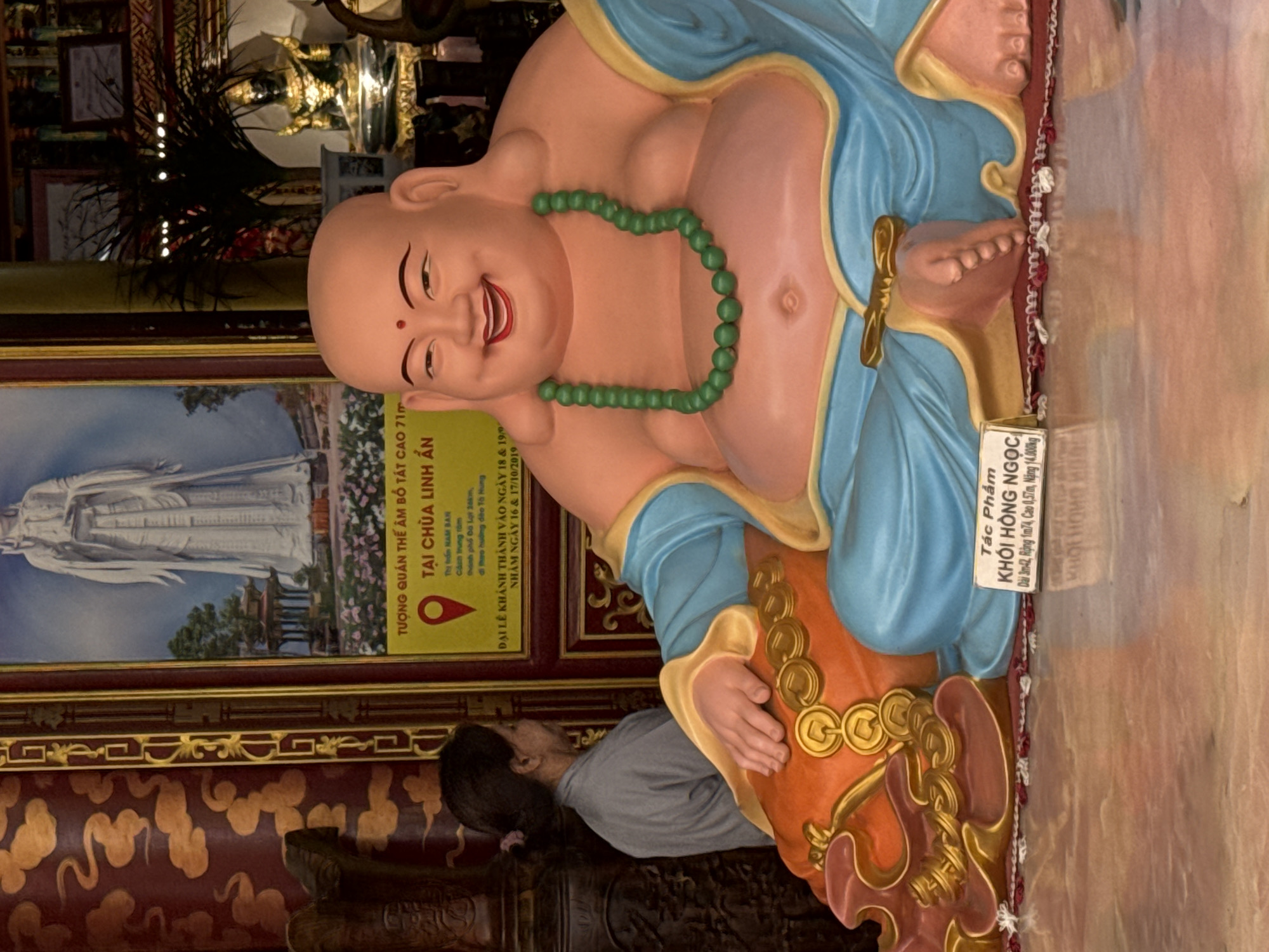 The image depicts a statue of a laughing Buddha, a common motif in Buddhist art, seated in a cross-legged position. The statue is adorned with a green beaded necklace and is draped in flowing blue robes. To the left of the statue, a woman in a grey garment is partially visible, facing away from the camera. Behind the Buddha, a large, framed painting shows a figure in white robes standing in a garden setting. A yellow sign with Vietnamese text is placed near the painting, indicating a location within the city of Xuân Thành, Vietnam, specifically mentioning "Chùa Linh An" which translates to Linh An Pagoda. The flooring appears to be polished stone with a reflective surface. The overall scene suggests an interior setting, likely within a temple or religious establishment.