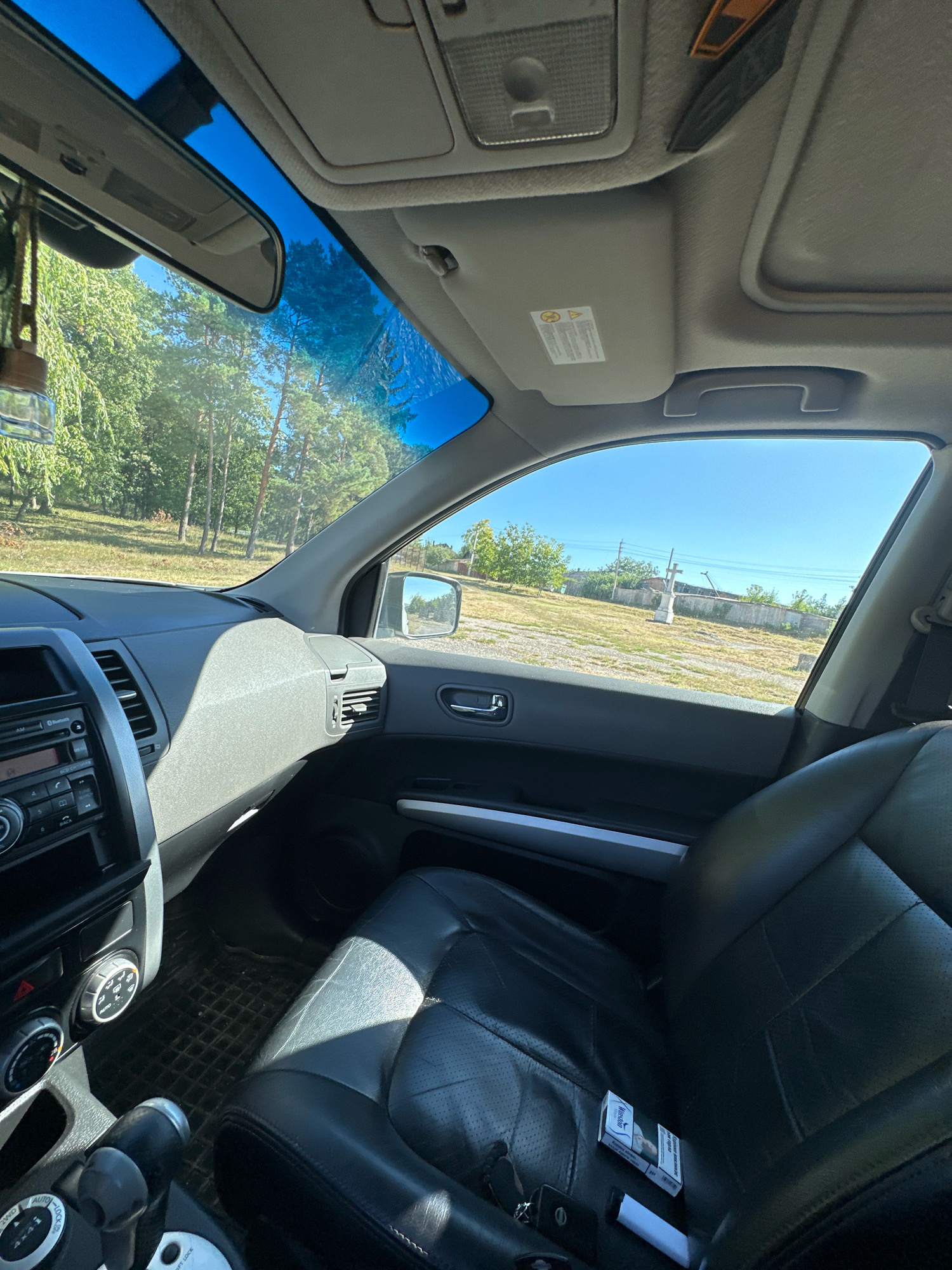The image shows the interior of a car, likely a Nissan X-Trail, parked outdoors on a sunny day. The view is from the passenger seat looking towards the driver's side and out the front and side windows. The car is parked on a dirt and gravel surface, with dry grass and trees visible outside. In the distance, across a field, there appears to be a small village with buildings and a stone structure with a cross. The car's interior features black leather seats, a dashboard with a radio and climate controls, a gear shifter, and a car key and a pack of cigarettes on the passenger seat. The overall impression is of a parked vehicle in a rural or semi-rural setting, possibly on the outskirts of Makiv, Ukraine, given the contextual information provided.