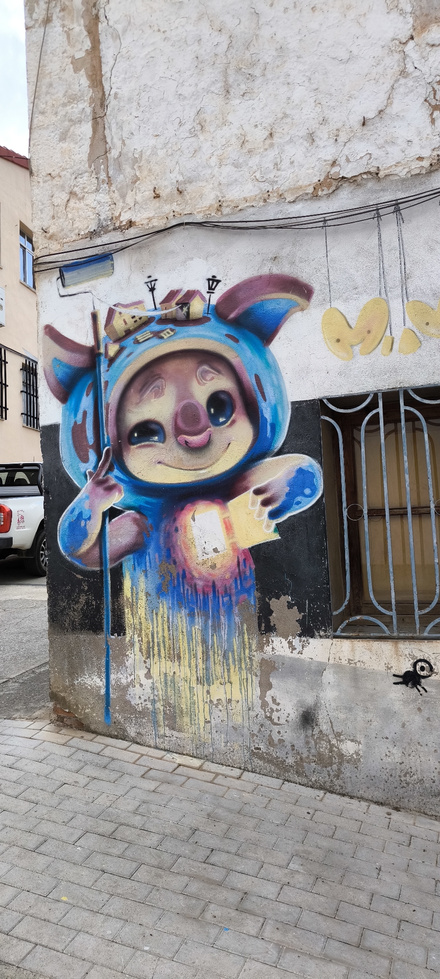 A vibrant mural of a cartoonish pig character is painted on the exterior wall of a building in Fanzara, Spain. The character, depicted with large ears and a blue hooded costume adorned with miniature houses and streetlights, is holding a paint roller and a can of paint. The mural extends down the wall, with colorful paint drips creating an abstract effect. A white pickup truck is visible parked to the left of the mural. The building's wall shows signs of age and wear, with peeling paint and cracks. A barred window is present on the right side of the mural. A small, stylized black creature is also painted on the wall near the bottom right corner. The ground in front of the wall is paved with light-colored bricks.