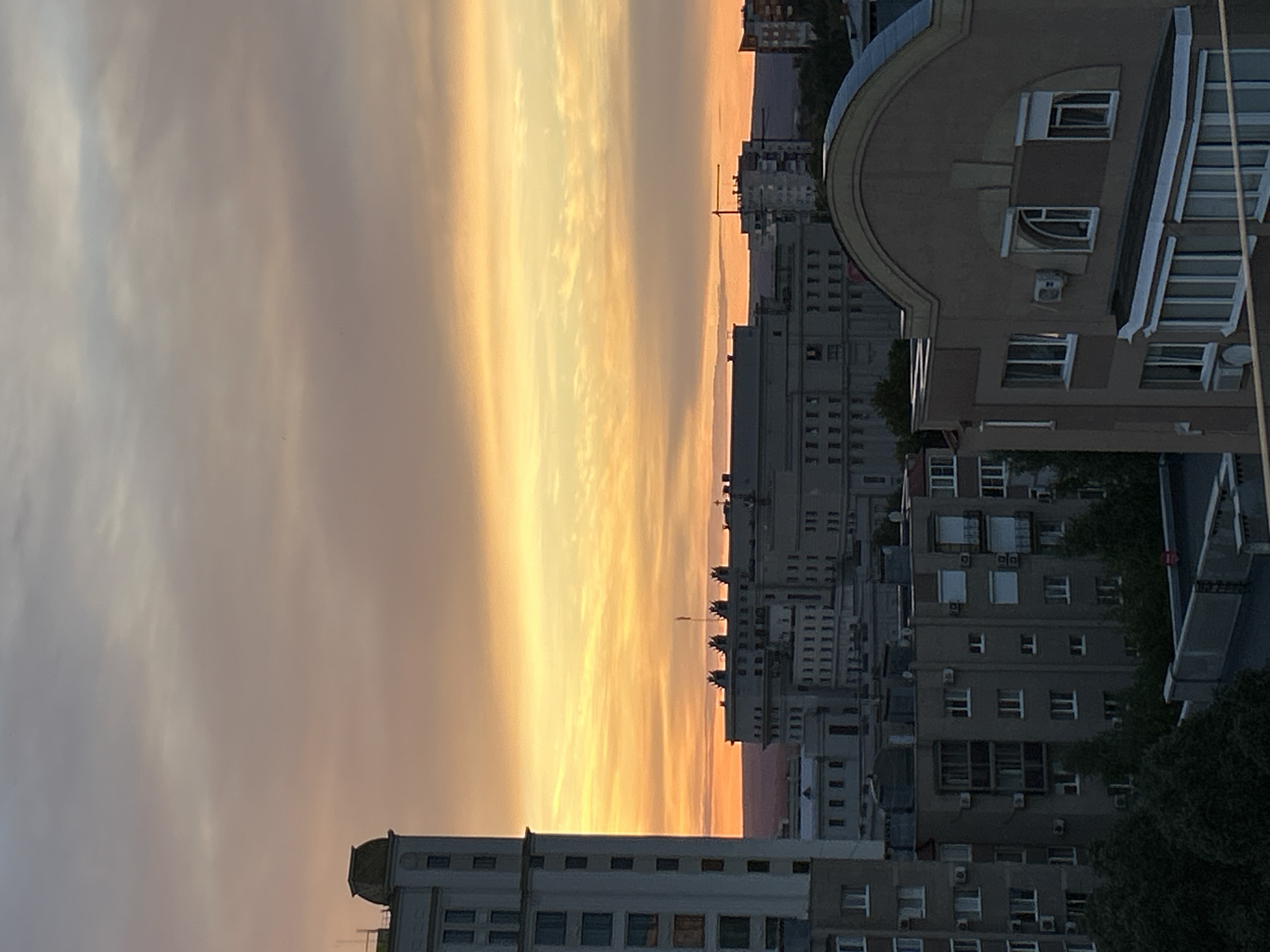 This is a vertical, outdoor, medium shot of a city skyline at sunset. The sky is filled with a gradient of soft oranges and yellows, with wispy clouds stretching across. To the right of the frame, several apartment buildings are visible. One building in the foreground has a distinctive curved facade. The buildings are predominantly grey and feature numerous windows and air conditioning units. Trees are sparsely scattered throughout the cityscape. The time of day is sunset, and the weather appears clear. The image depicts a residential urban setting. No people or text are visible. The location is Novokuybyshevsk, Russia.