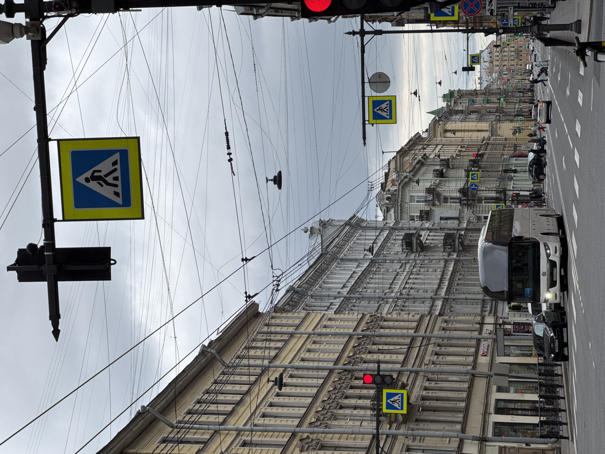 This street-level, eye-level, color photograph depicts a street scene in Saint Petersburg, Russia, characterized by dense overhead wiring and pedestrian crossings. The orientation is vertically oriented.
Buildings line both sides of the street. The architecture features ornate facades with repetitive window patterns. The buildings are light beige.
Overhead, a dense network of wires spans the street, connecting to poles. The sky is overcast.
Traffic is present, with a white bus and a black car visible. Pedestrians are crossing the street. Traffic lights are mounted on poles alongside pedestrian crossing signs. The signs are blue and yellow.
The street is paved and marked with zebra crossings. The general atmosphere is urban.