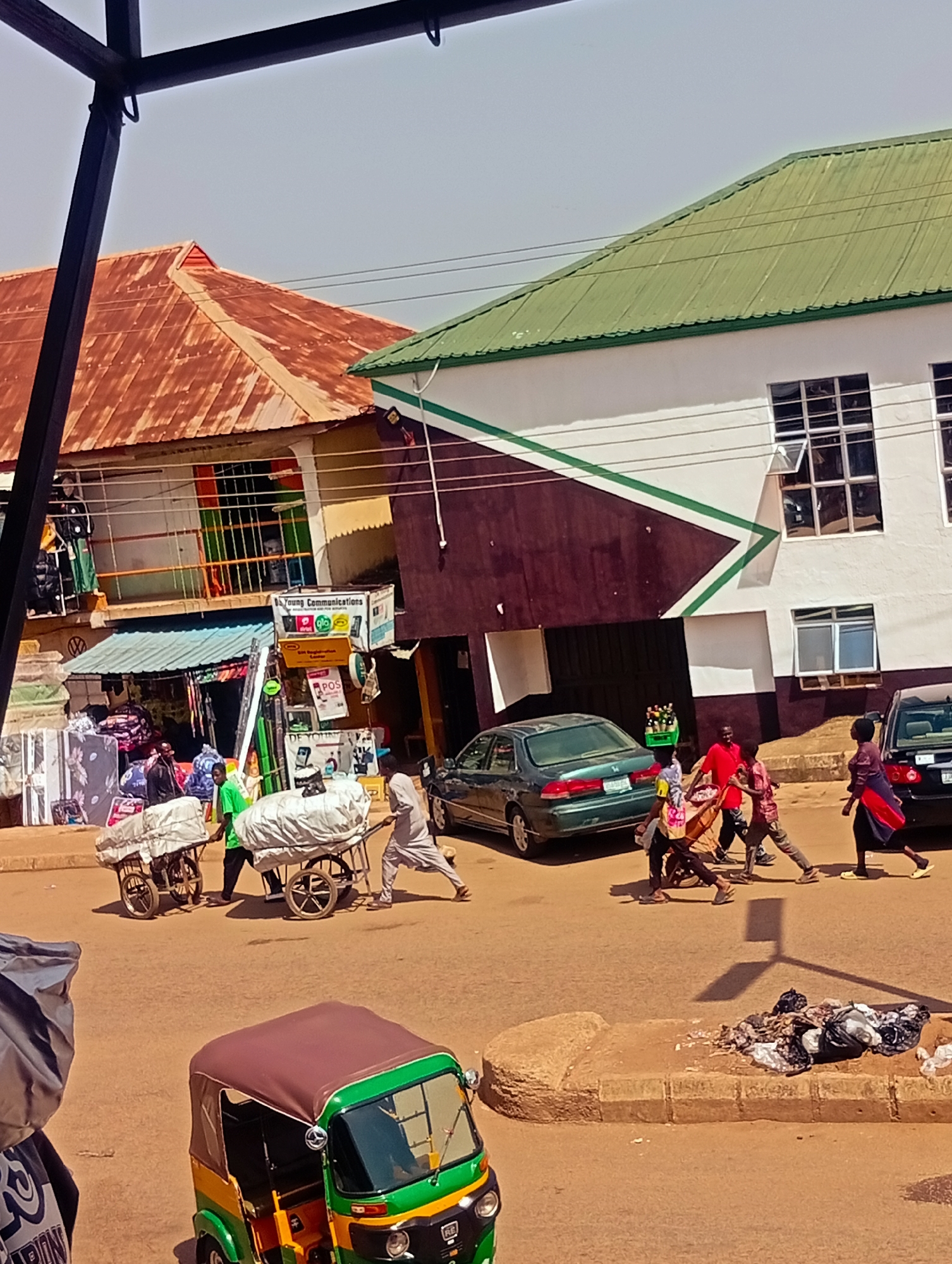 The image depicts a busy street scene in Bukuru, Nigeria. In the foreground, a green and yellow auto-rickshaw is parked. Beyond the auto-rickshaw, two men are pushing carts laden with goods wrapped in white fabric. To the right, a group of people are walking, including two individuals carrying a large object. A green sedan is parked on the street, and further down, another dark car is visible.
The buildings in the background have corrugated metal roofs, one rusty orange and the other green. The building with the green roof is painted white with a dark maroon section and green diagonal stripes. Several windows with metal bars are visible on the buildings. A sign hanging from one of the buildings reads "Young Communications" and advertises "Glo SIM Registration Center". Various items are displayed outside the shops, including what appears to be mattresses. A pile of garbage bags is on the curb. The ground is paved with asphalt. The sky is a clear, pale blue. The overall impression is a vibrant, commercial area with activity.