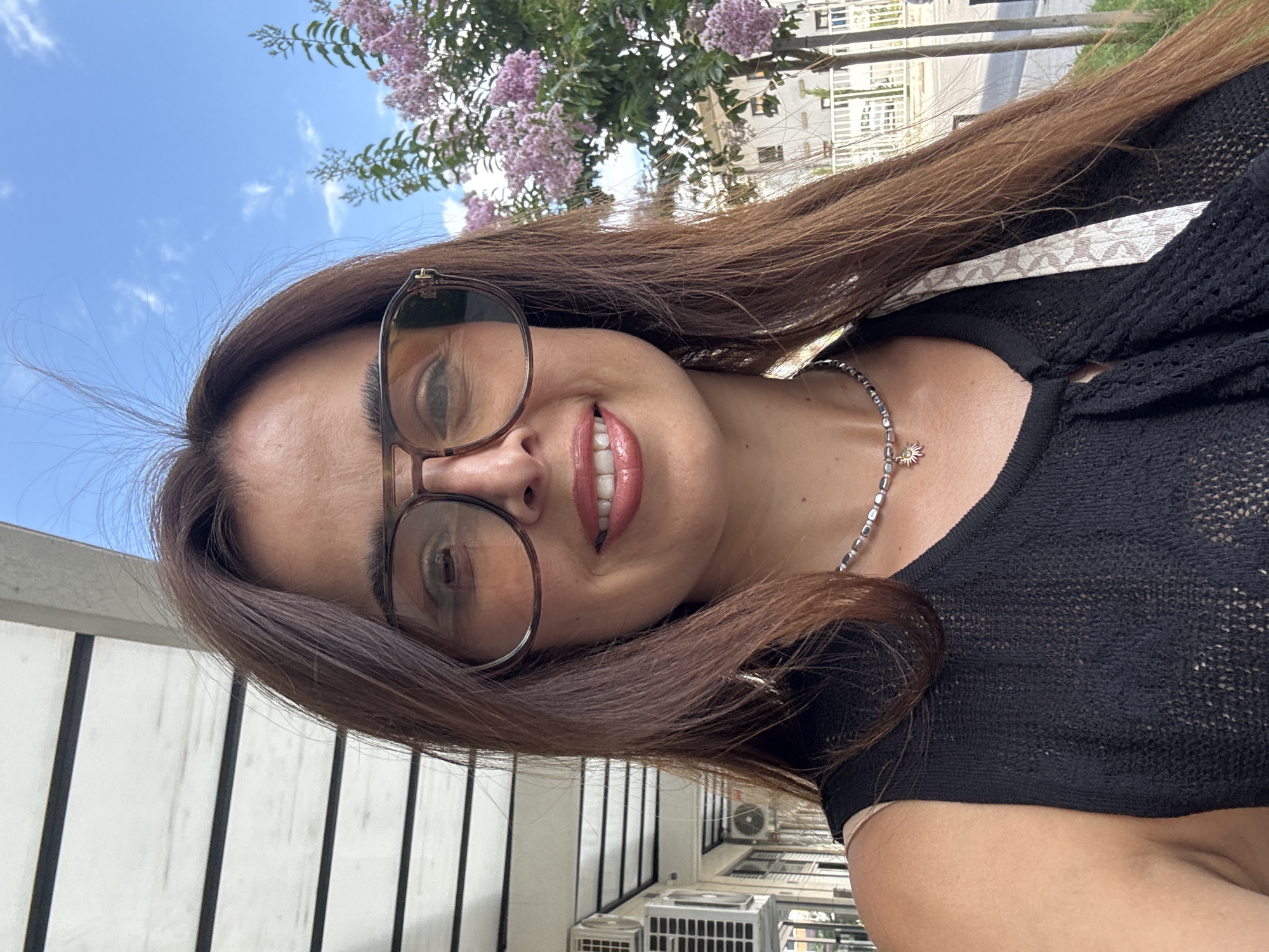 A medium shot captures a woman taking a selfie outdoors, with a bright blue sky and wispy clouds overhead. She has long, dark brown hair that falls past her shoulders and is wearing large, amber-tinted glasses with thick brown frames. Her lips are painted a glossy pink, and she has a slight smile. She is dressed in a black, sleeveless, textured top and wears a delicate silver necklace with a small sun pendant. A portion of a beige strap with a repeating pattern is visible across her shoulder and chest.
The background suggests an urban setting. To her left, there is a white structure with horizontal black accents, likely an awning or architectural feature, and an air conditioning unit is visible attached to the wall. To her right, a tree with clusters of small pink flowers is in bloom. A white fence and some buildings can be seen further in the distance. The lighting indicates it is daytime, with the sun casting subtle shadows.