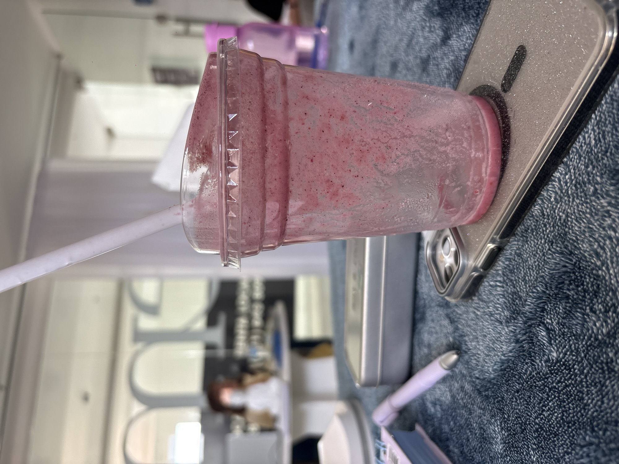 A partially consumed red smoothie drink in a clear plastic cup with a white straw is resting on a silver phone and a light blue, fluffy surface. A light purple pen is nearby. The background shows a blurred interior of a building with white walls, furniture, and equipment, including chairs and medical-related machinery. A person in a white coat and face mask is partially visible. This location is Puebla, Mexico.