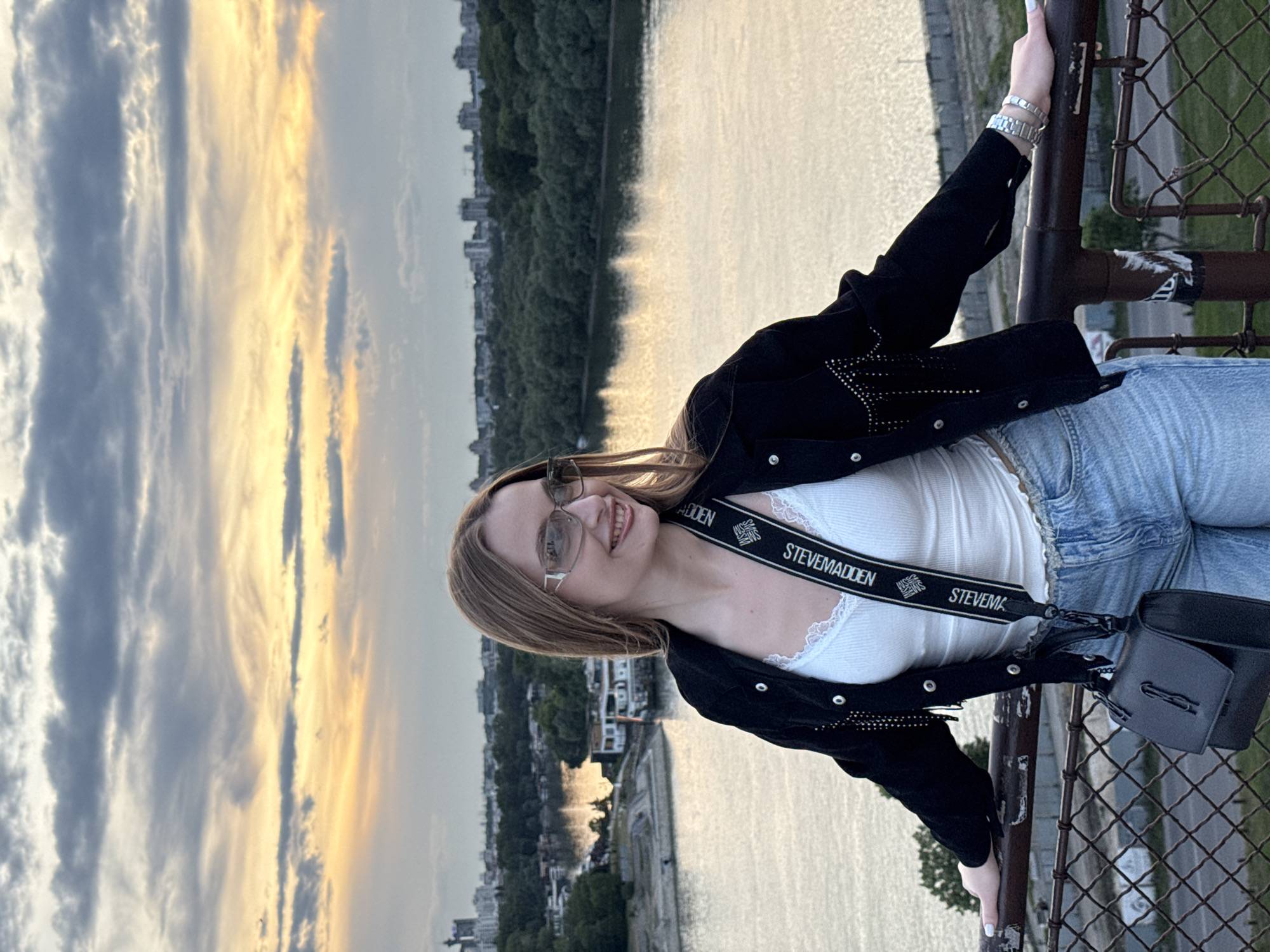 A young woman stands on a bridge overlooking the Sava River in Belgrade, Serbia. She is leaning against a metal fence. The landscape features a river, vegetation, buildings, and a sunset sky. The woman is wearing a black jacket with embellishments, a white top with lace trim, light-wash jeans, and sunglasses. She has a black purse with a "Steve Madden" strap. A city skyline and sky filled with clouds are visible in the background.