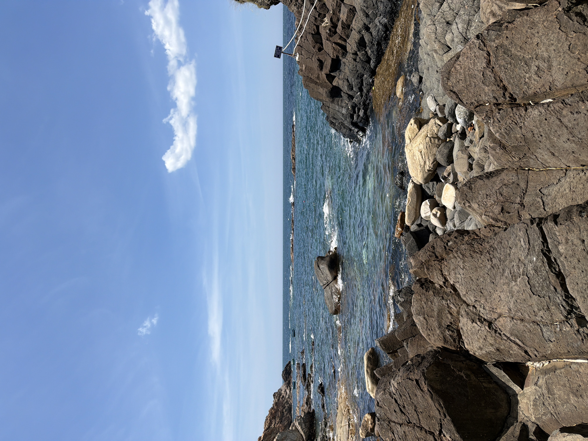 The image shows a rocky coastline on a clear day. The ocean is a deep blue with waves breaking against the shore. There are several large rocks in the foreground and mid-ground, some of which are partially submerged in the water. In the distance, the ocean stretches to the horizon, meeting a bright blue sky with a few white clouds. A small signpost is visible on a rock ledge on the right side of the image, with white ropes leading away from it. The scene appears to be a natural coastal landscape, possibly a public area given the signpost and ropes, suggesting it might be a designated viewpoint or a safe access point for visitors. The general impression is one of a tranquil and rugged natural environment. The location is identified as Phú Lương, Vietnam, a coastal region known for its scenic beauty.