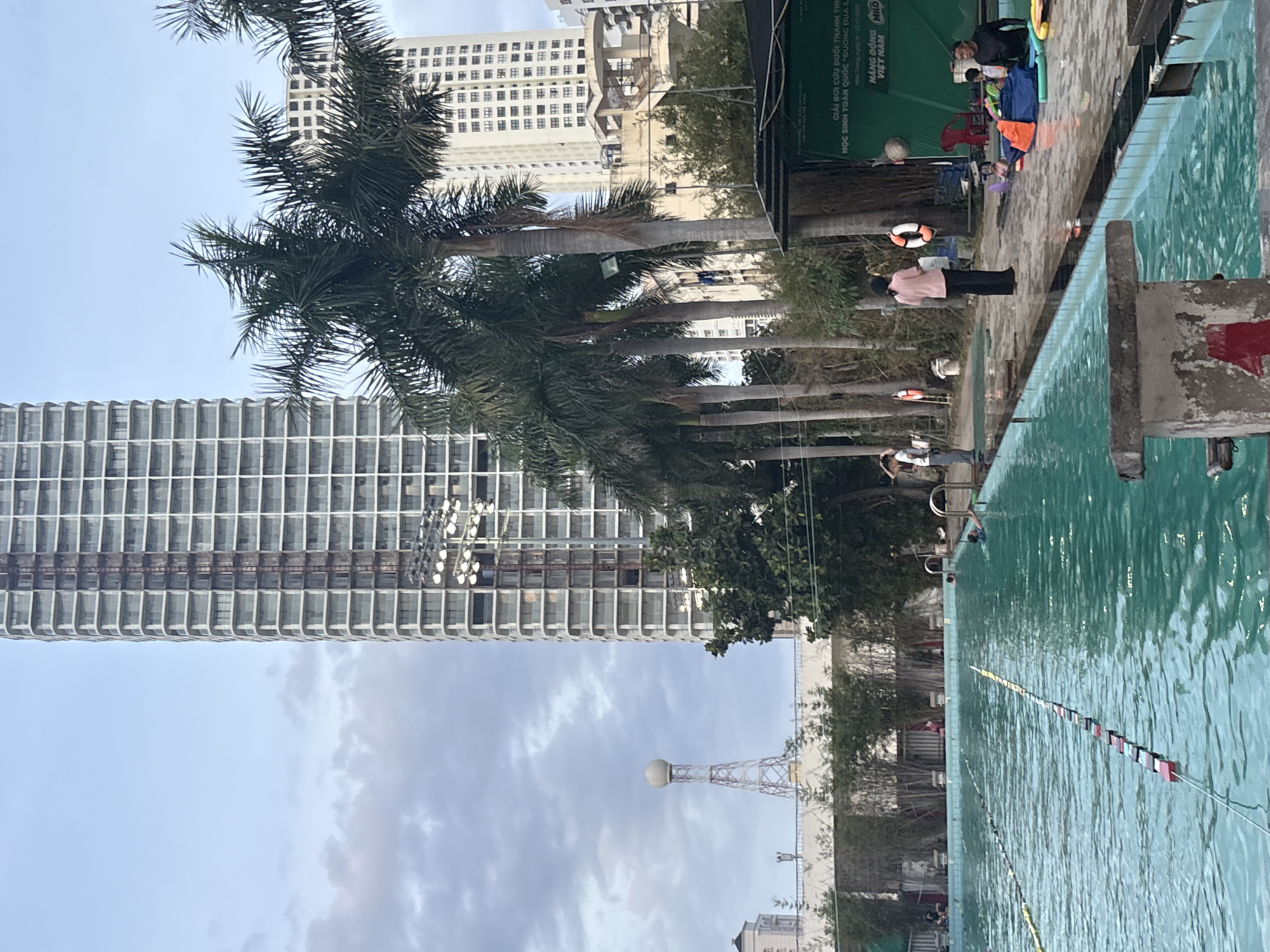 The image depicts an outdoor swimming pool area in Nha Trang Ward, Vietnam. In the background, a tall, modern apartment building dominates the skyline, with construction visible on its upper floors. To the left of the apartment building, a water tower with a spherical top is present. Several palm trees are scattered throughout the scene, adding a tropical element to the urban environment.
The swimming pool itself is large and rectangular, with lanes marked by buoys. The water is a clear turquoise color. Several people are visible around the pool. In the water, a few individuals are swimming, including one person in a blue swimsuit and another in a darker swimsuit. On the poolside, a woman in a pink shirt and dark pants is standing near a life preserver. Another person, possibly an instructor, is standing further away near a metal railing. A group of children are gathered near the edge of the pool, some with colorful flotation devices.
A black structure, possibly a kiosk or building, is situated at the far end of the pool area, with a sign on it that appears to be in Vietnamese. The lighting suggests it is either early morning or late afternoon. The sky is partly cloudy, with a blend of blue and gray hues. The overall impression is one of a recreational facility within a bustling city.