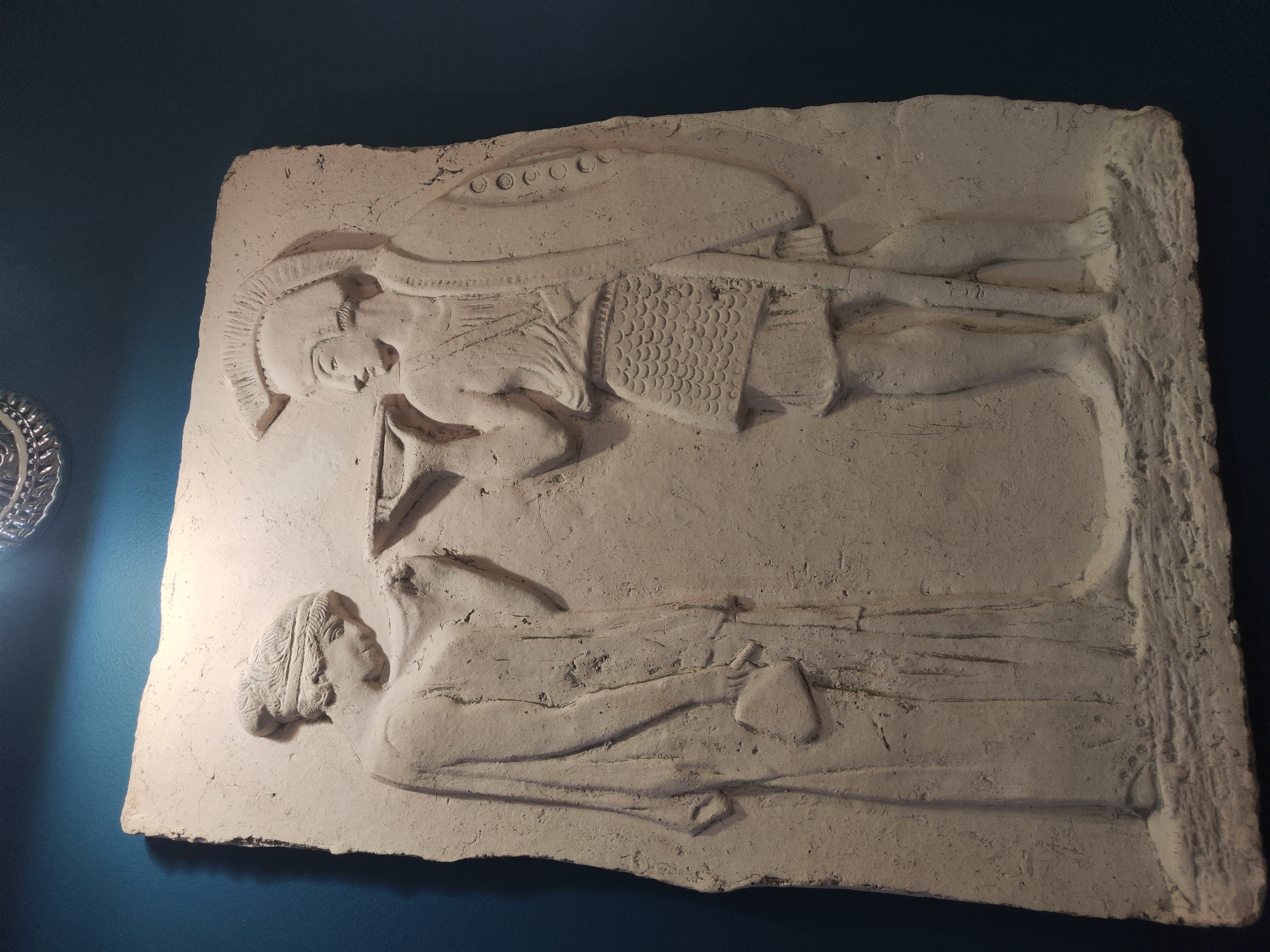 This is a close-up, angled photograph of a pale, bas-relief stone carving mounted on a dark blue wall. The carving depicts two figures. On the left is a woman with her head bowed, wearing a long, draped garment and a headband. She holds a small, book-like object in her left hand. On the right is a male figure in armor, wearing a Corinthian helmet, a chest plate resembling scales, and holding a round shield with circular decorations on his left arm. He is offering a shallow bowl with his right hand to the woman. The carving has a rough, textured surface and appears to be an ancient artifact. The overall lighting suggests an indoor museum or gallery setting, with a bright light source above and to the left casting shadows. This relief is part of the collection of the State Hermitage Museum in Saint Petersburg, Russia.