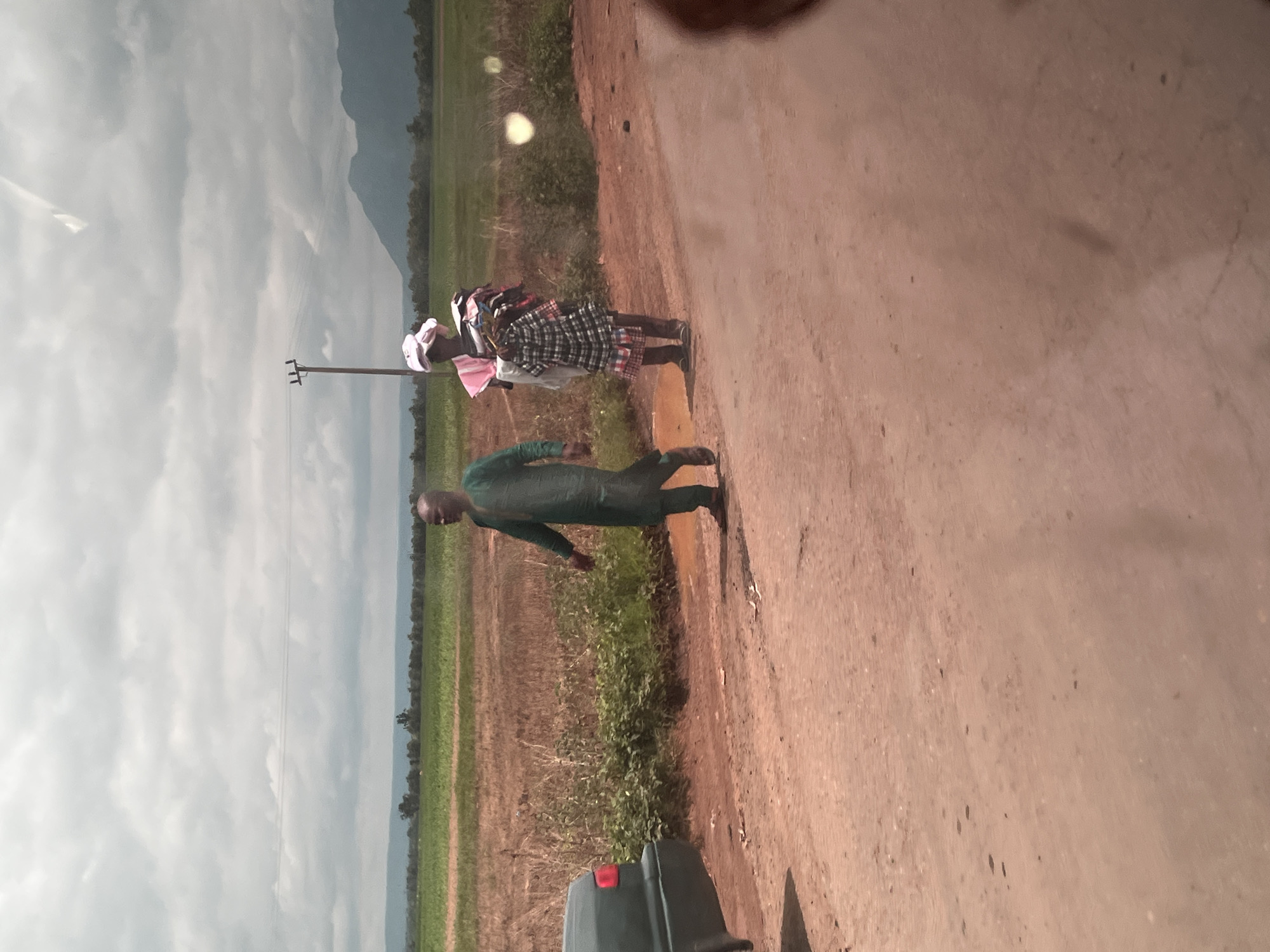 In Karshi, Nigeria, on a dirt road, two individuals are visible. A man wearing a green traditional outfit is walking away from the viewer, his right arm extended. To his right, a person wearing a pink headscarf and a checkered dress carries a bundle of clothing. Behind them, a sparse field of dry grass and green crops stretches towards a distant mountain range under a cloudy sky. A utility pole stands in the middle ground. A dark green car is parked on the left side of the road.