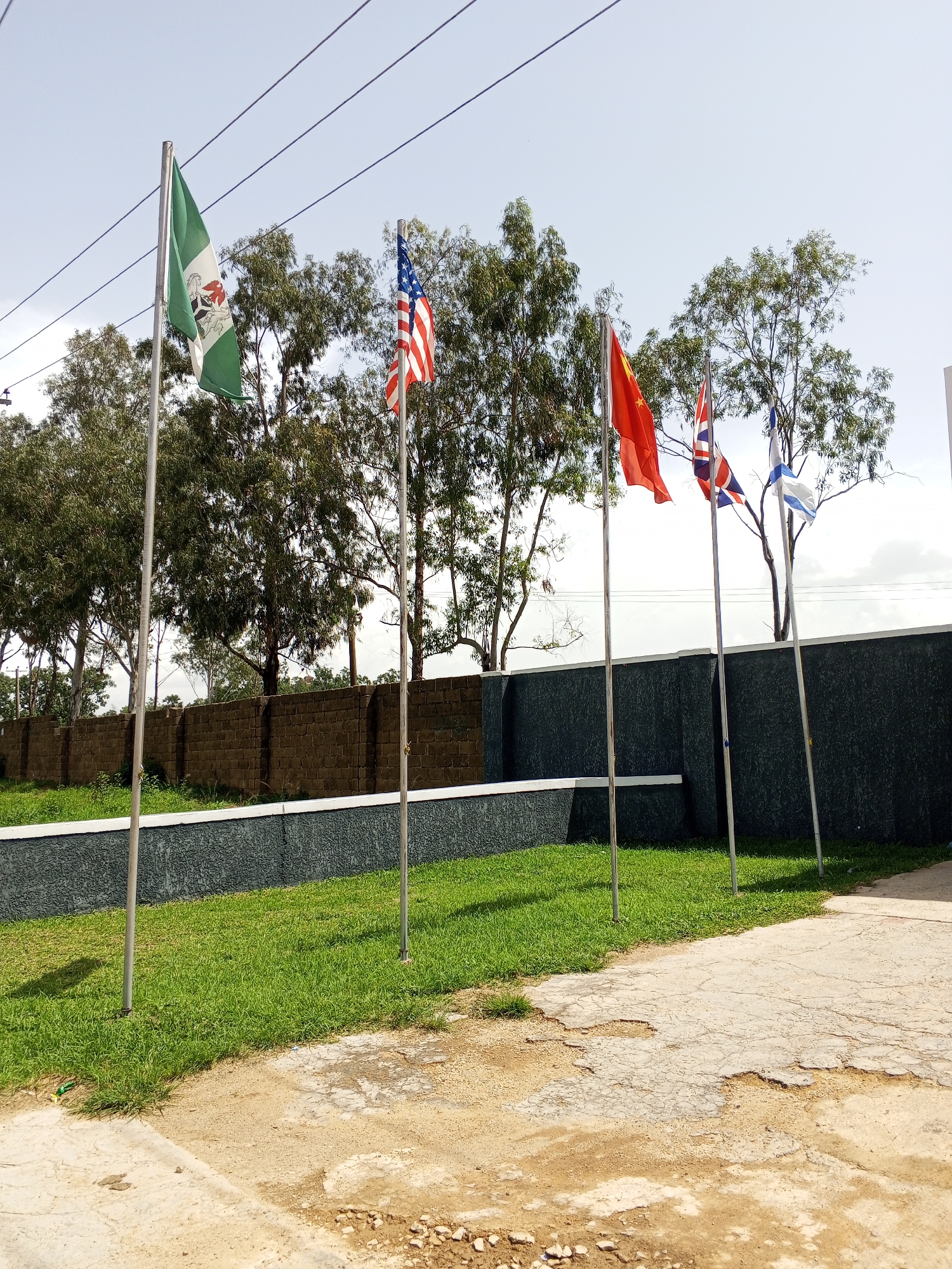 In Bukuru, Nigeria, an outdoor scene features five flagpoles arranged in a line against a backdrop of trees and a brick wall. The flags are, from left to right: Nigeria, the United States, China, Great Britain, and Israel. The flags are in various states of unfurling, suggesting a gentle breeze. The ground in the foreground is a mix of concrete and dirt, with patches of dry, cracked concrete. A grassy area with a white-bordered dark wall separates the flags from the brick wall behind them. The sky is clear and pale blue. There are no people or animals visible in the image.