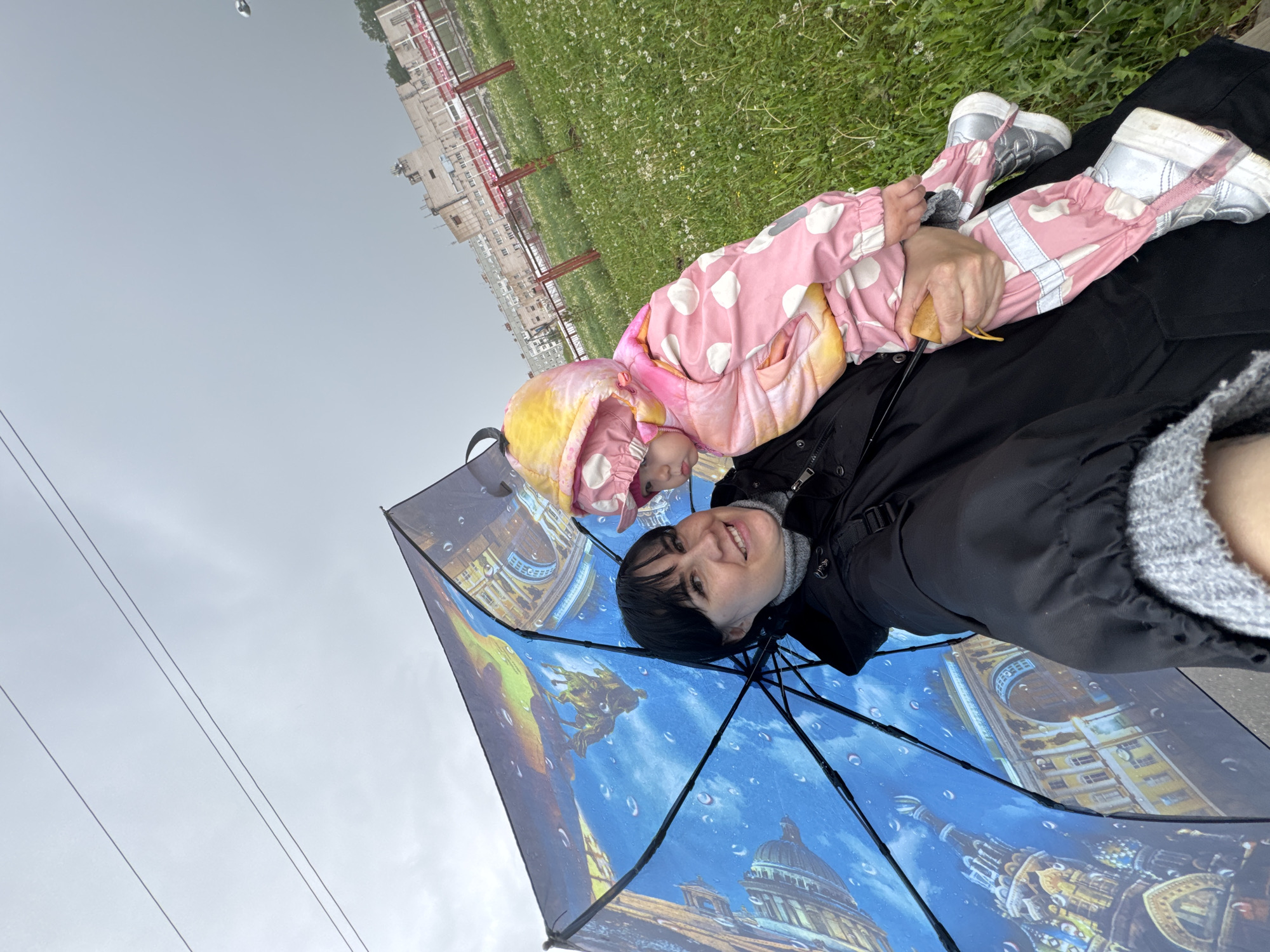A woman is lying on her back, holding a young child in a pink and white snowsuit on top of her. The woman is holding a large umbrella with a blue sky and building images on it. The background features green grass, a building in the distance, and a gray sky. Overhead electrical wires are visible in the upper left corner. It is not possible to conclusively determine that this photo was taken in Zav'yalovo, Russia.