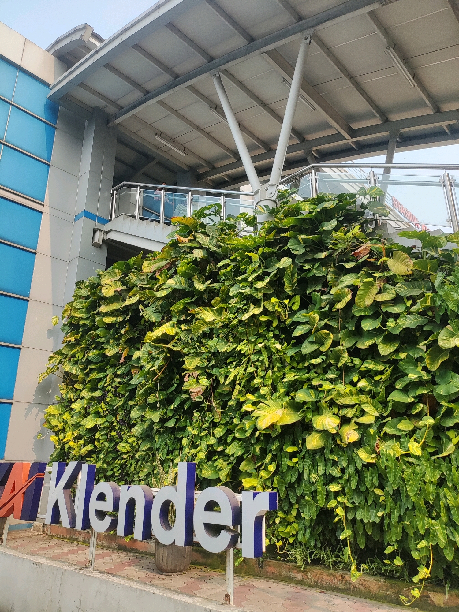 The image shows an outdoor scene with a building featuring blue glass panels and a metal canopy supported by white pillars. A lush green wall of foliage, possibly a vertical garden or overgrown plants, covers a significant portion of the lower building facade. In front of the foliage, large white and blue 3D letters spell out "Klender," indicating a location name. The ground in front of the sign is paved with patterned tiles. In the background, a ramp or staircase with a glass and metal railing is visible leading up to the building. The sky is clear and bright, suggesting daytime. This scene is located at Klender Station in East Jakarta, Indonesia, a public transportation hub.
