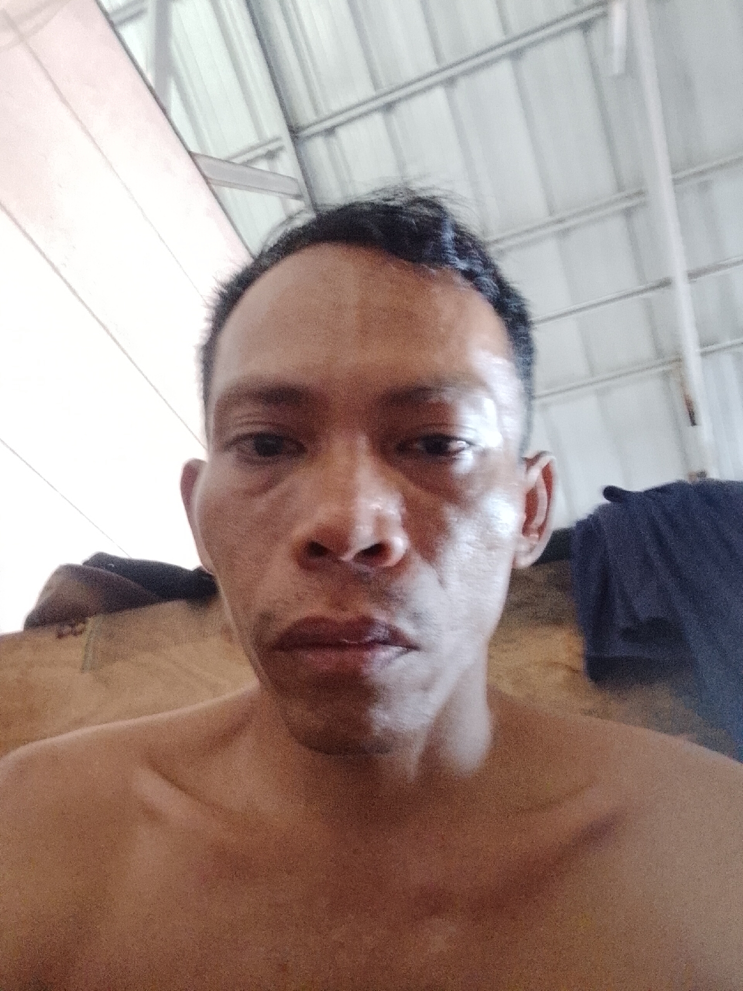 A selfie of a shirtless, middle-aged Asian man with short, dark hair in an indoor environment. He has a neutral expression. A section of a corrugated metal ceiling is visible in the background. A dark blue cloth hangs in the upper right corner, and a tan, textured surface is visible behind the man's left shoulder. The location is Bukit Ketok, Indonesia.