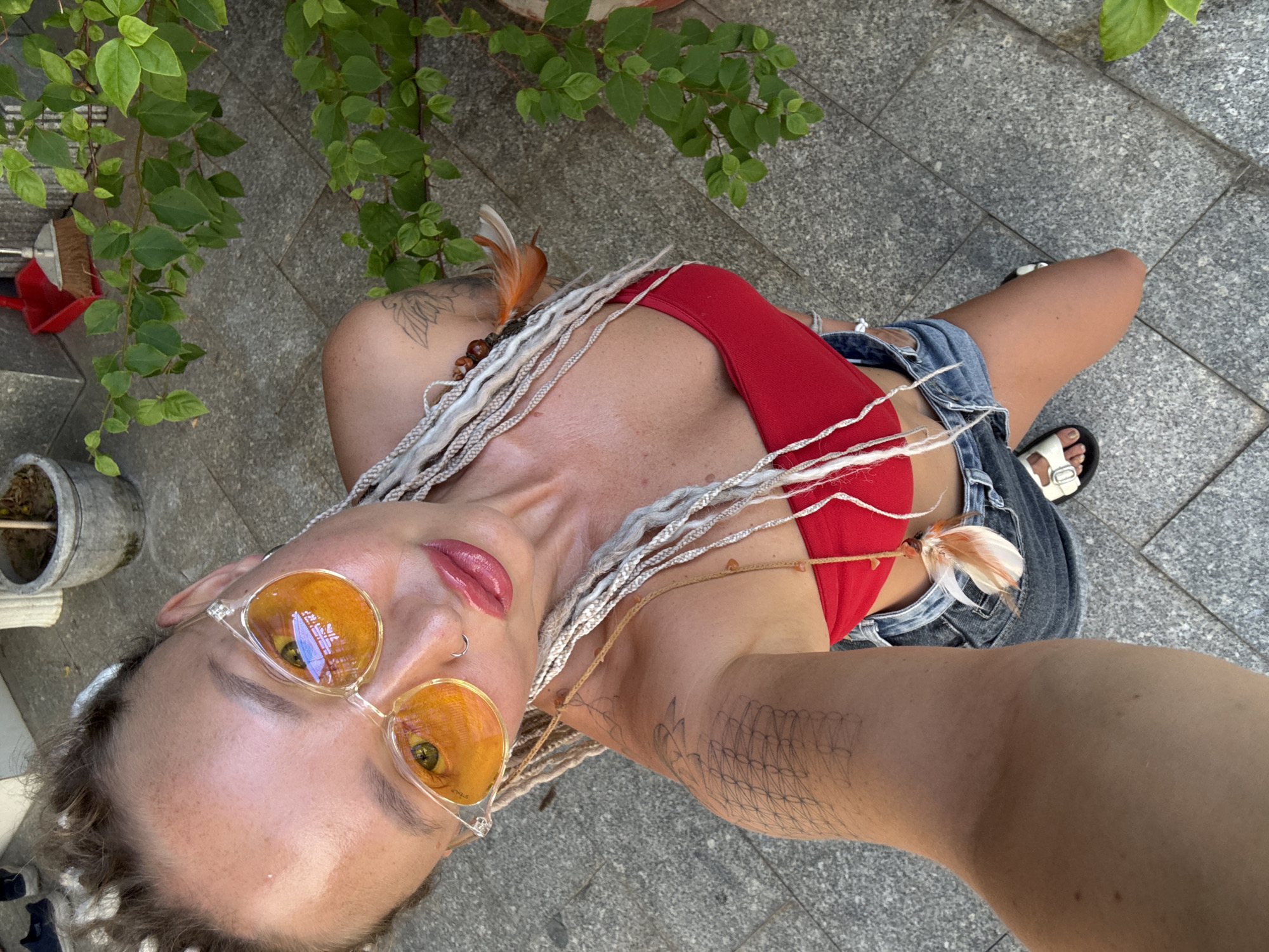 A young adult with white braided hair, wearing sunglasses, a red top, and denim shorts, is posing for a selfie outdoors. The background suggests a paved area, possibly a sidewalk or patio, with some greenery and potted plants visible. The scene appears to be set in a city environment, consistent with an urban setting like Nha Trang, Vietnam. The individual is the primary focus, with the surroundings providing context.