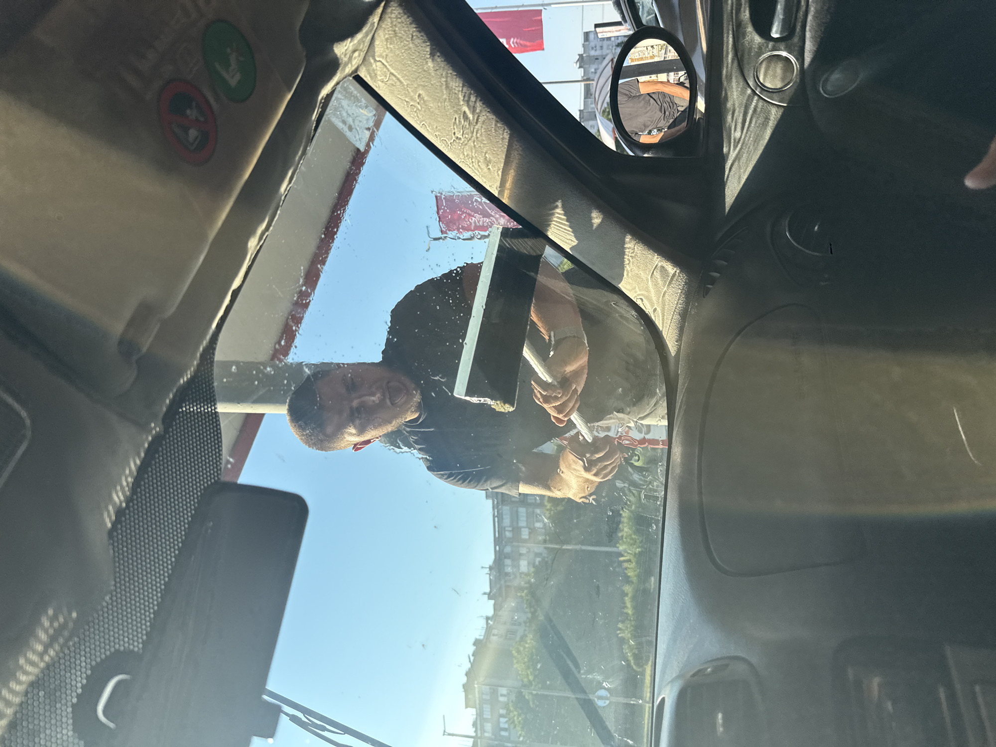 An interior view from a car in Burgas, Bulgaria, shows a man cleaning the windshield from the outside with a squeegee. The man, wearing a dark t-shirt, is positioned in front of the car, his face partially obscured by the windshield and sunlight. Water droplets are visible on the glass. In the background, buildings and trees suggest an urban environment. The car's interior, including the dashboard, rearview mirror, and side mirror, is partially visible. The side mirror reflects another person, also wearing a dark t-shirt. The scene is illuminated by bright sunlight.