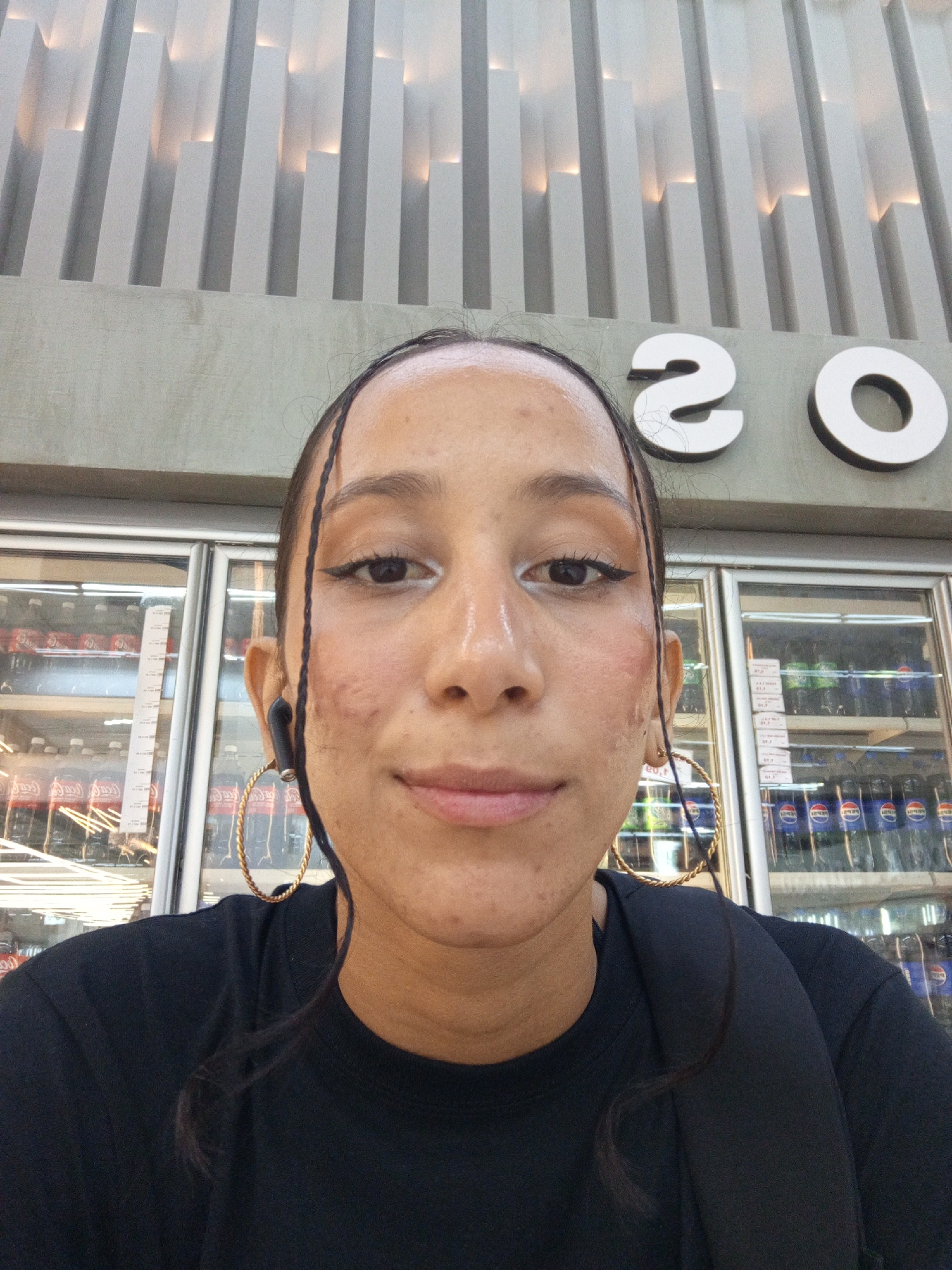 A medium-shot selfie shows a person with brown skin, dark eyes, and thin eyebrows, wearing a black t-shirt and large gold hoop earrings. The person has thin strands of hair framing their face. They are also wearing black earbuds in their left ear. Behind the person is a commercial refrigerated display case stocked with soft drinks. Above the display case, a sign reads "20". The background also includes a gray, textured architectural wall feature with vertical white accents. The location is likely a storefront or commercial establishment in Porlamar, Venezuela.