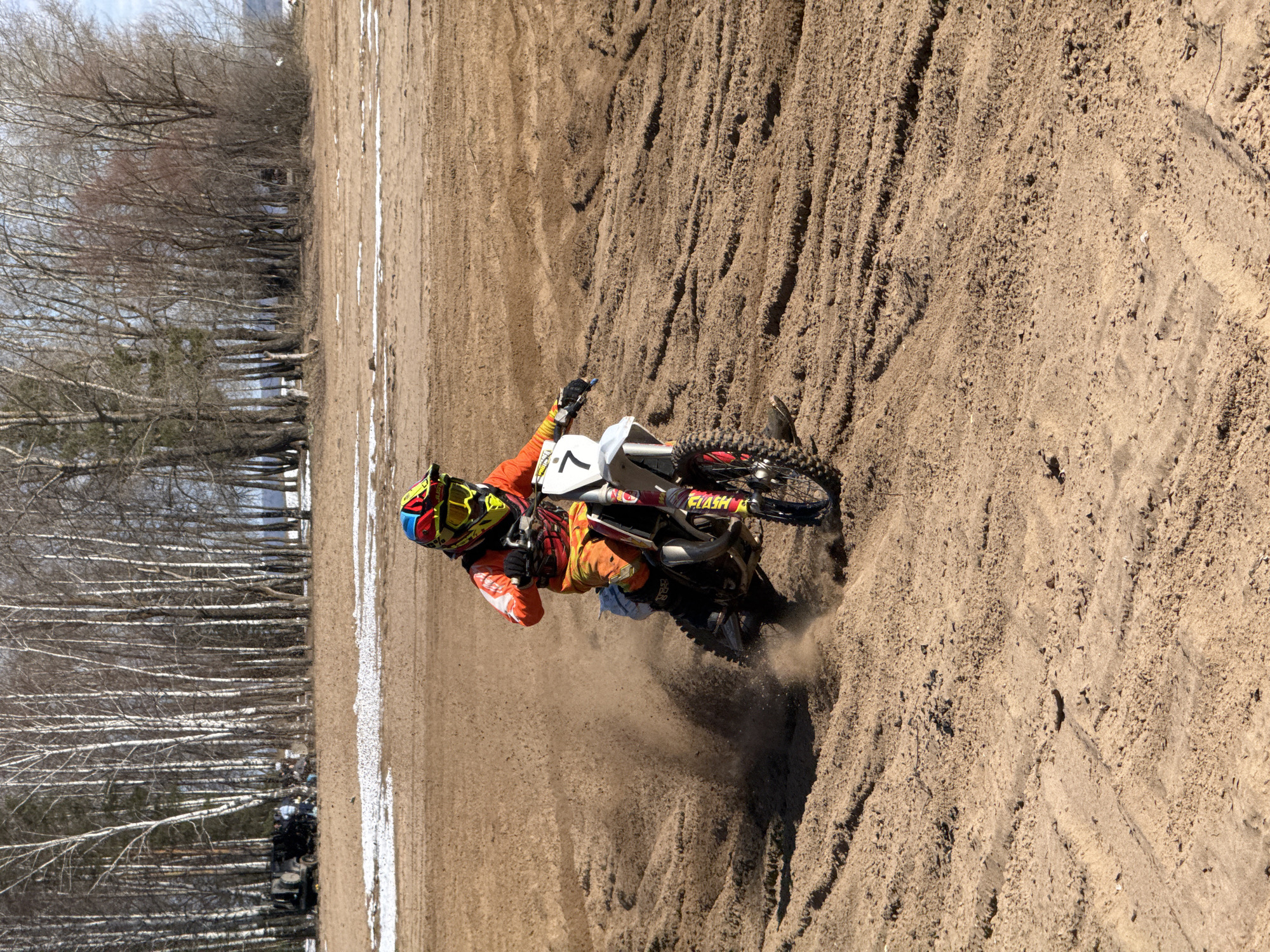 A dirt biker wearing an orange and black suit, protective helmet, and gloves rides a motorcycle on a dirt track. The motorcycle has the number "7" on its side and a sticker on the rear fender. The track is sandy and rutted, with trees lining the side. The photo was taken in Noviy, Russia.