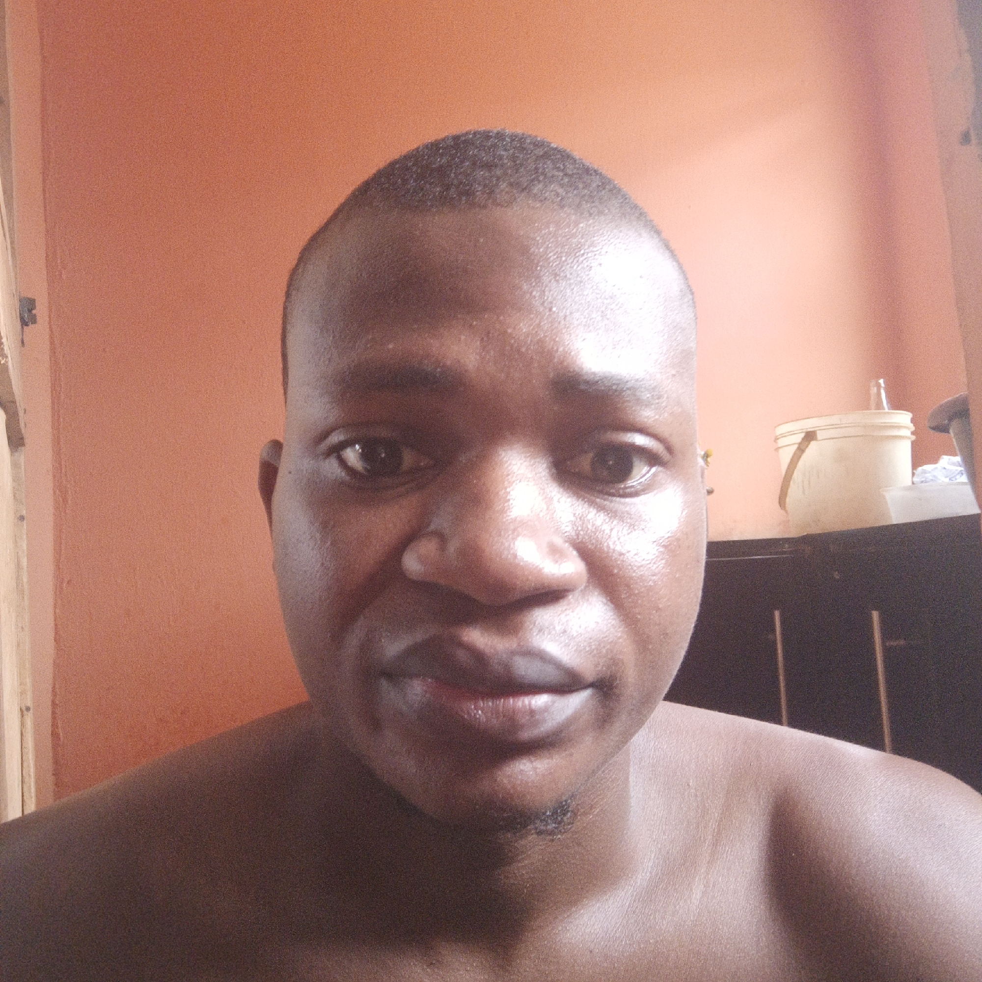 A close-up, front-facing photograph features a dark-skinned man with a shaved head. He appears to be shirtless. The background consists of a light orange wall. To the right, there is a dark-colored cabinet or furniture with a white bucket placed on top. Given the prompt, the location is presumed to be Akwanga, Nigeria.