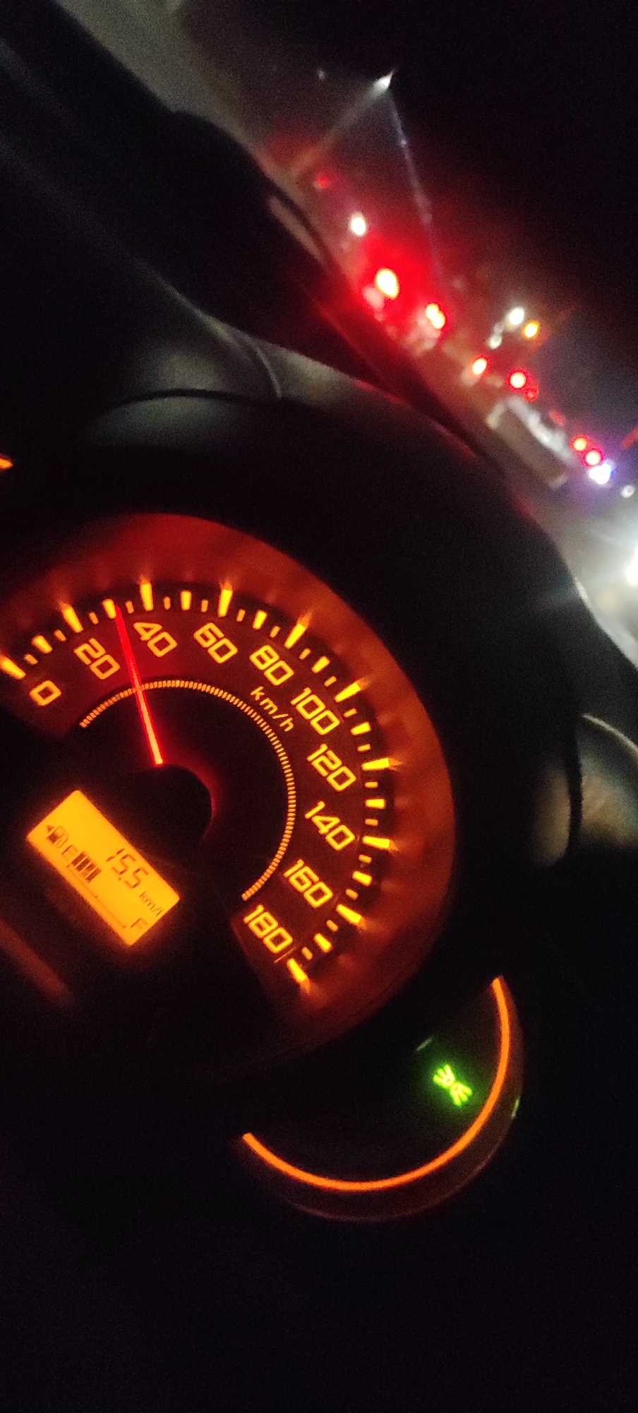 The image shows a close-up view of a vehicle's illuminated dashboard at night. The speedometer needle points between 20 and 40 km/h, specifically at approximately 25 km/h. A digital display shows "15.5 km/l," likely indicating fuel consumption. The dashboard is predominantly orange and red, with markings for speed in km/h. In the background, blurry red and white lights suggest other vehicles or traffic signals, consistent with a street scene in Bolo Krajan, Indonesia. There are no people visible, and the focus is solely on the vehicle's internal display. The overall environment appears to be a road at night.