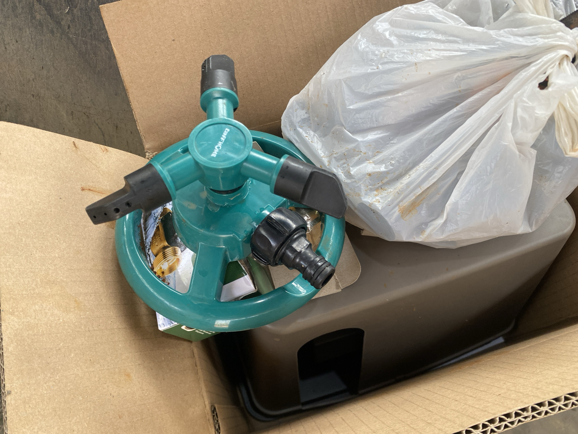 An overhead shot shows a new, teal-colored, three-arm oscillating sprinkler sitting in a cardboard box. The sprinkler has a black nozzle connector and black sprayer tips. A white plastic bag, partially filled with dirt or compost, is to the right of the sprinkler. A tan-colored plastic tub or container is visible beneath the sprinkler and bag. The background consists of the brown interior of the cardboard box. The location is unknown.
